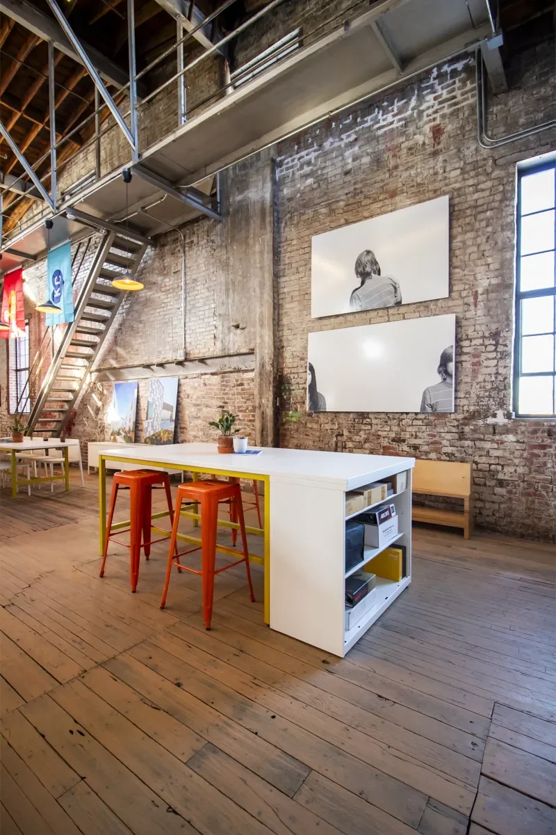 Industrial loft office with exposed brick walls, wooden floors, mezzanine staircase, white worktable, and orange stools