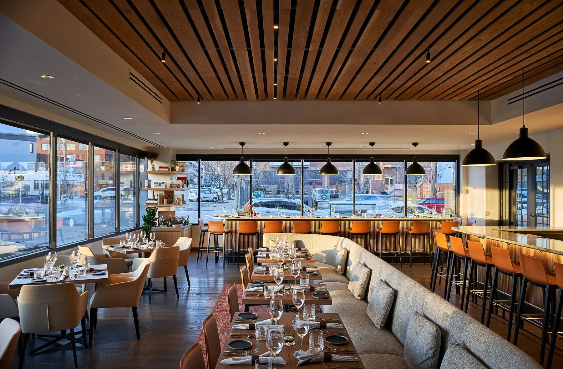 Modern upscale restaurant interior with set dining tables, orange bar stools, pendant lights, and floor-to-ceiling windows