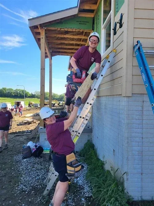 Two volunteers in safety helmets working on a house exterior, carrying a wooden board up a ladder at a construction site.