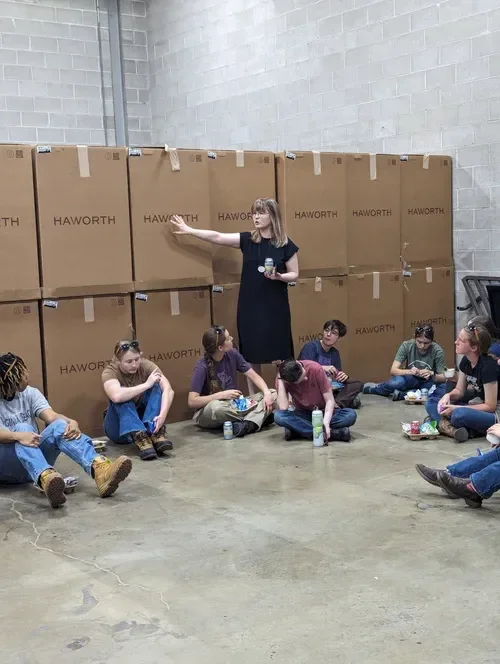 Woman leading a seated group discussion in a warehouse surrounded by stacked Haworth boxes