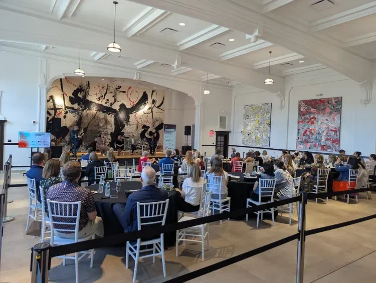 Art event in a bright gallery with seated attendees, tables, stage performers, and large abstract paintings.