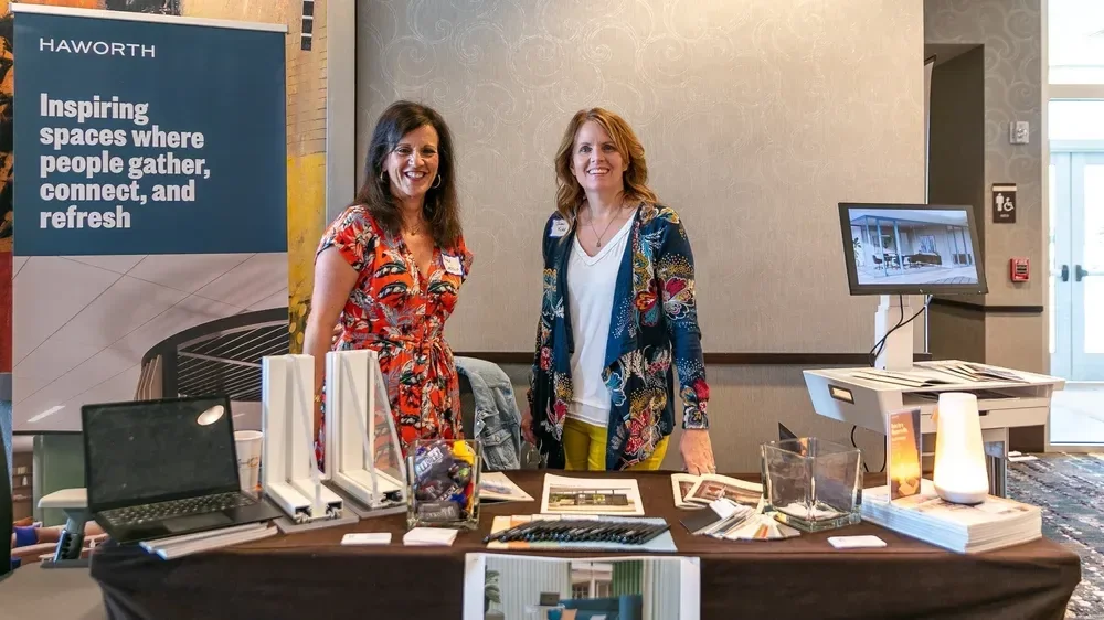 Two women stand behind a Haworth office furniture display table at an event.