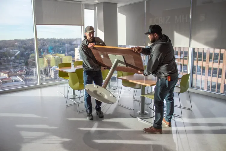 Two workers move a large tabletop through a modern office lounge with floor-to-ceiling windows.
