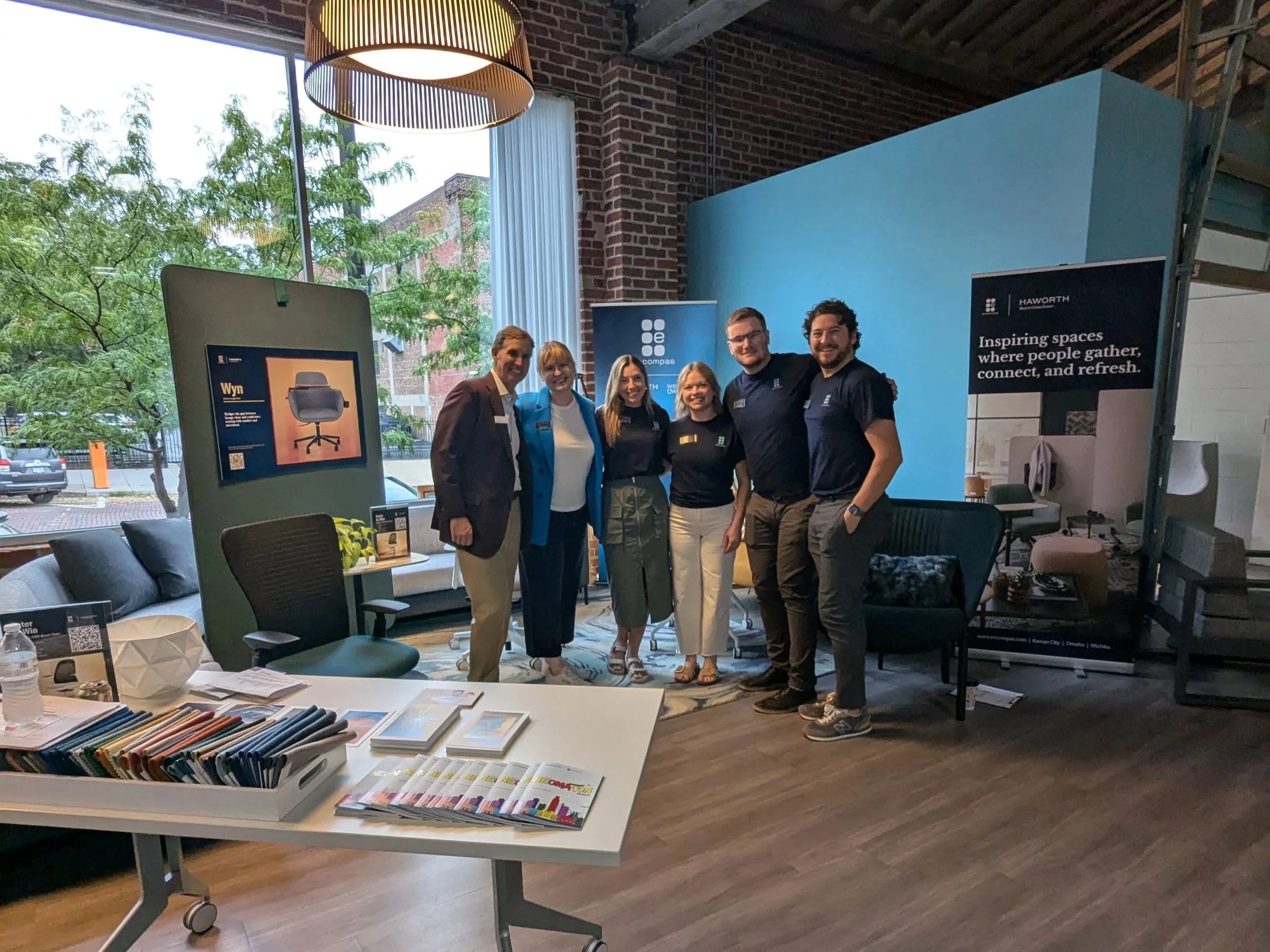 Six people pose in a modern office furniture showroom beside Haworth and Compass branding displays.