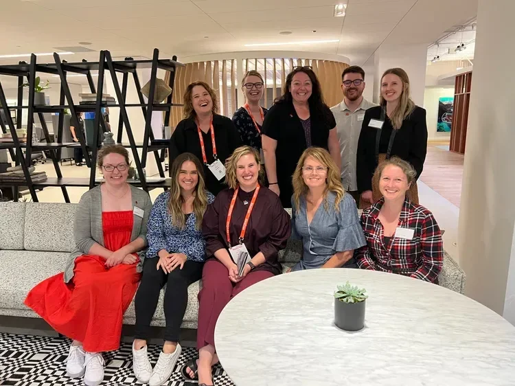 Group of nine smiling coworkers posing in a modern office lounge around a table with a potted succulent.