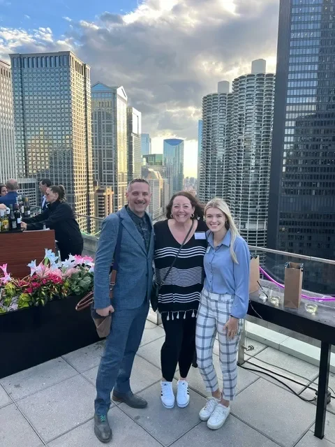 Three people pose on a rooftop terrace overlooking a downtown city skyline.