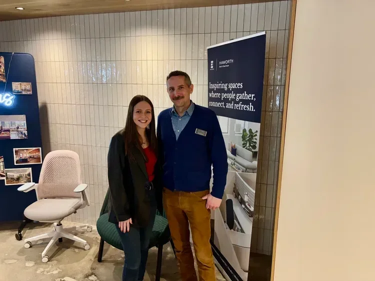 Two people pose beside a Haworth office furniture display featuring ergonomic chairs and a workplace design sign.