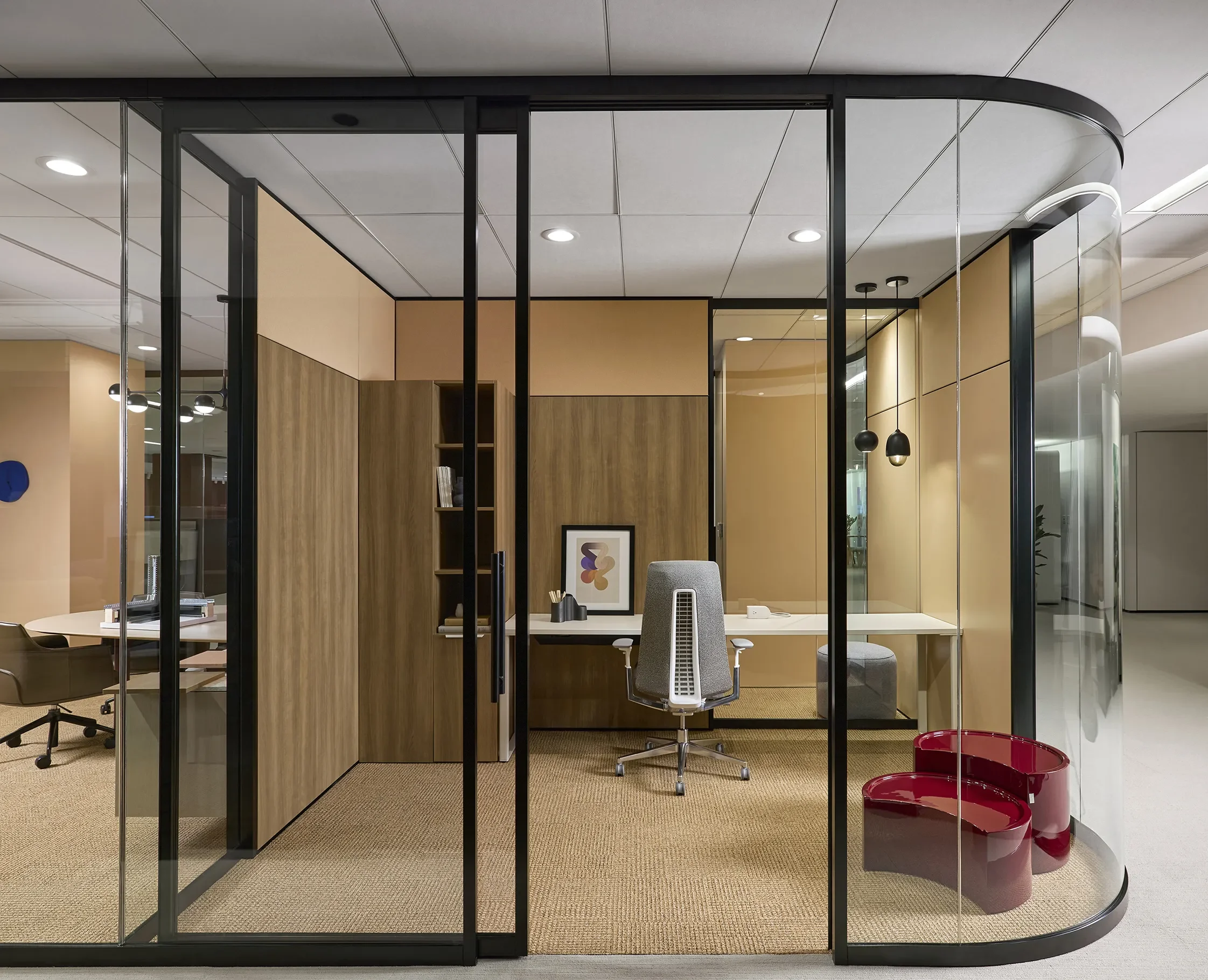 Modern glass-walled office with wood cabinetry, desk, ergonomic chair, pendant lights, and red lounge chairs