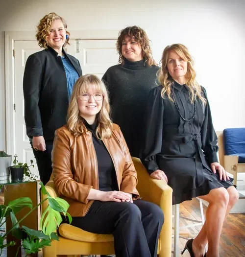 Four smiling women posing together indoors, with one seated in a mustard-yellow chair and houseplants nearby