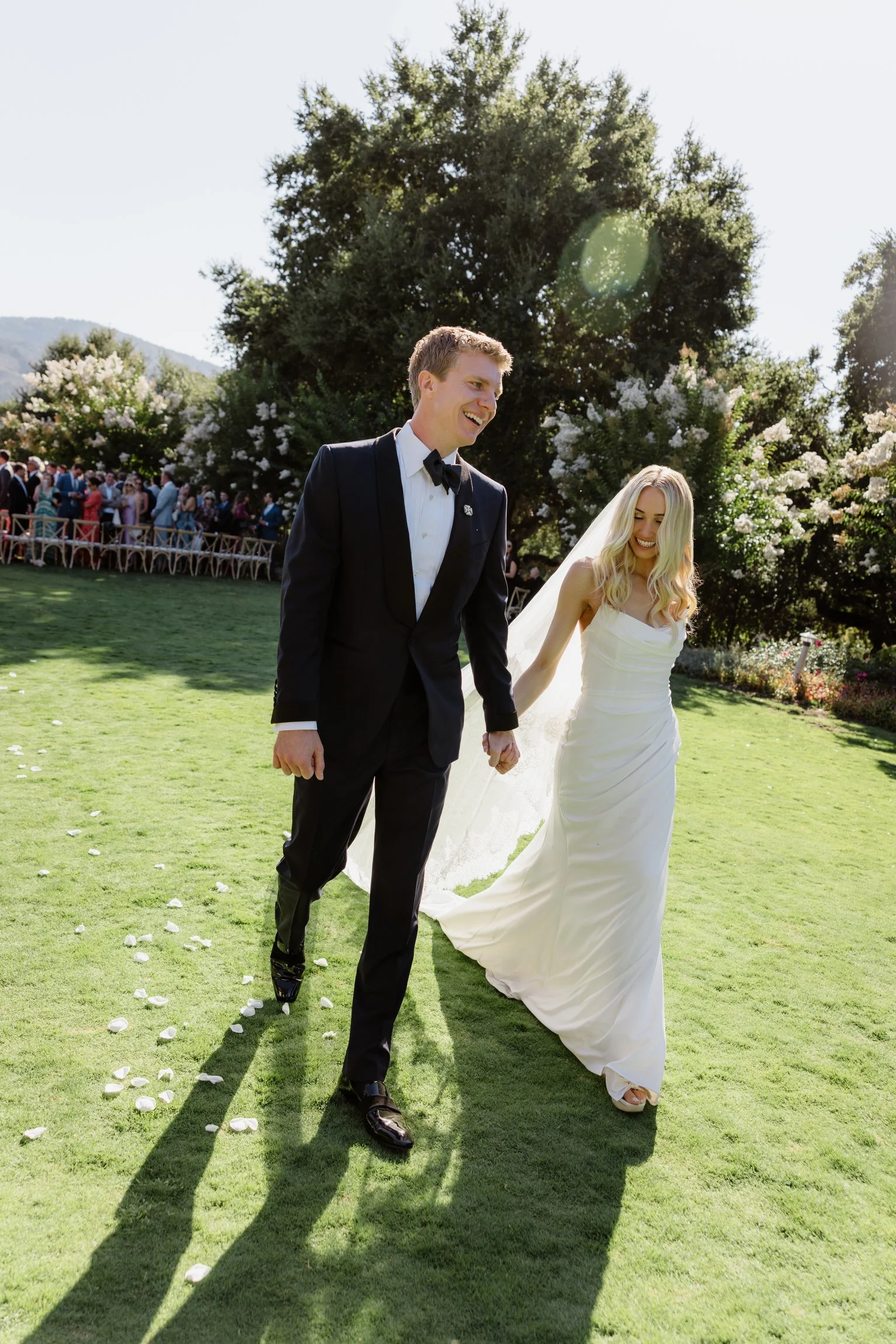 Happy bride and groom walking hand in hand on their wedding day on a sunny outdoor lawn with guests and trees in the background.