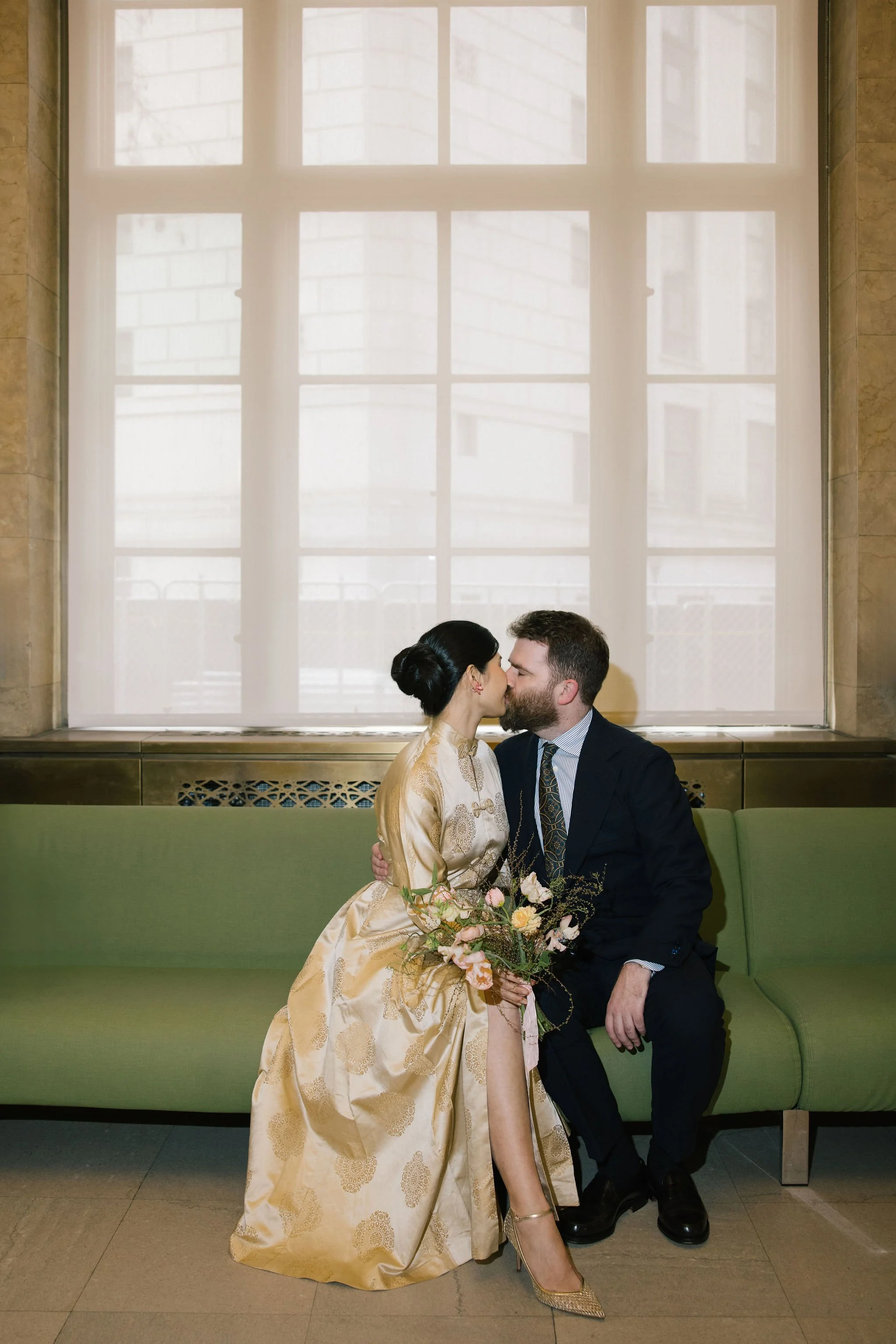 A man and woman, dressed in formal attire, share a kiss while seated on a green bench with a large window behind them. The woman wears a gold gown and high heels, holding a bouquet of flowers, and the man wears a dark suit and tie. The window is framed by beige stone walls.