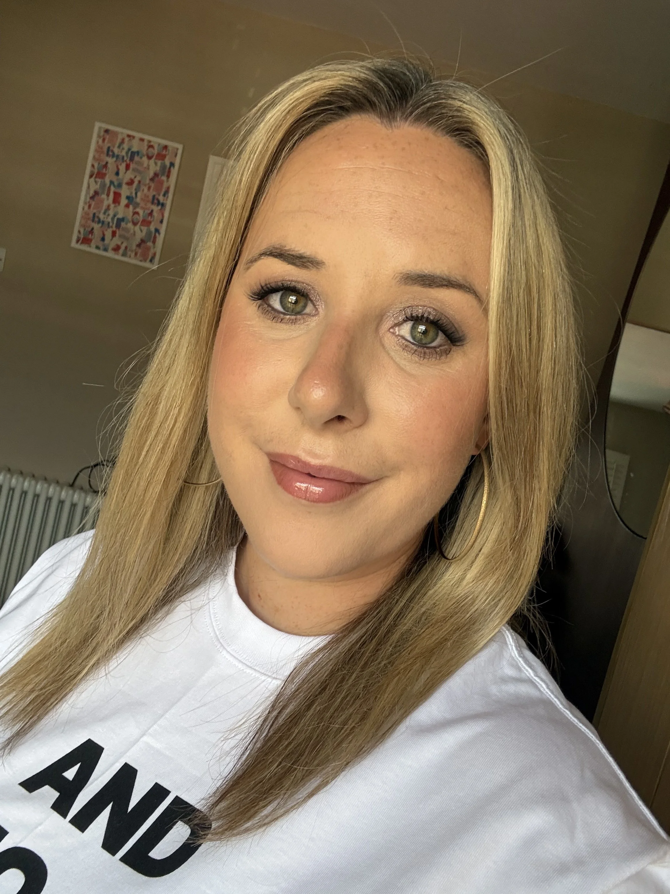 Close-up of a woman with blonde hair, hazel eyes, wearing makeup, gold hoop earrings, and a white shirt with black text.