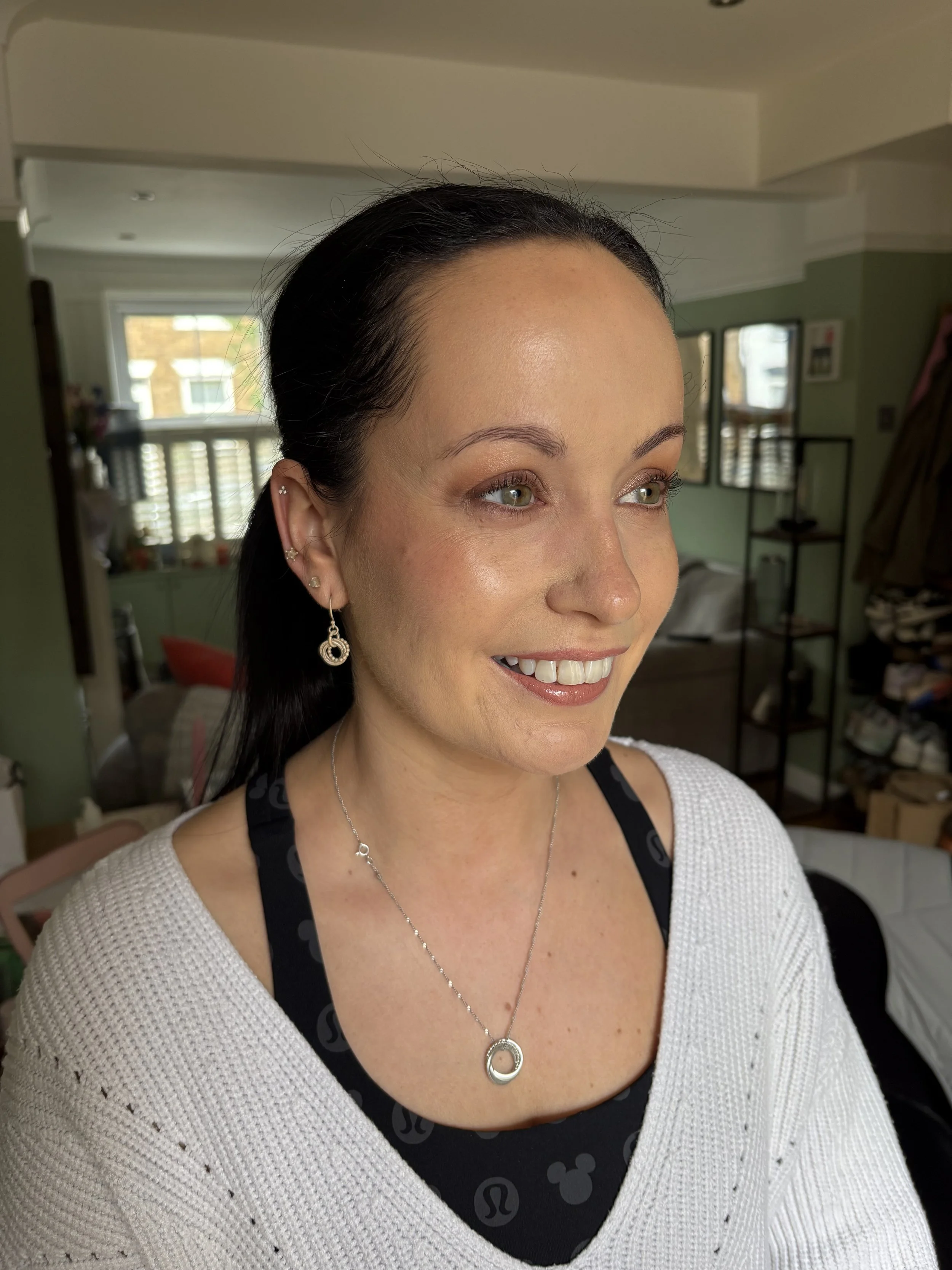 Close-up of a smiling woman with dark hair, wearing earrings and a necklace, inside a room with shelves and windows.