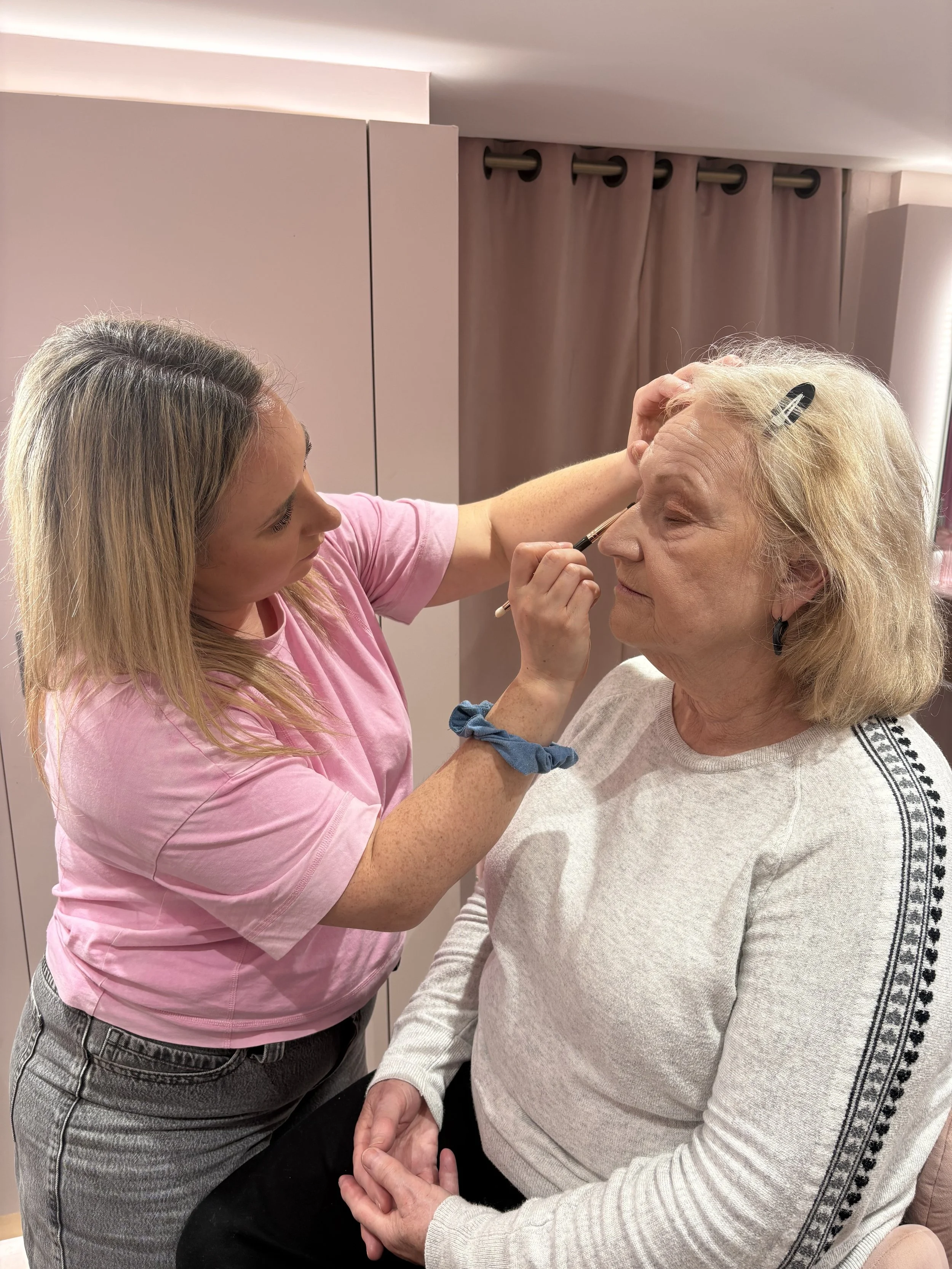 A woman with blonde hair is getting her eyebrows done by a beautician in a room with pink and beige walls. The beautician is holding a small brush near the woman's eyebrows.