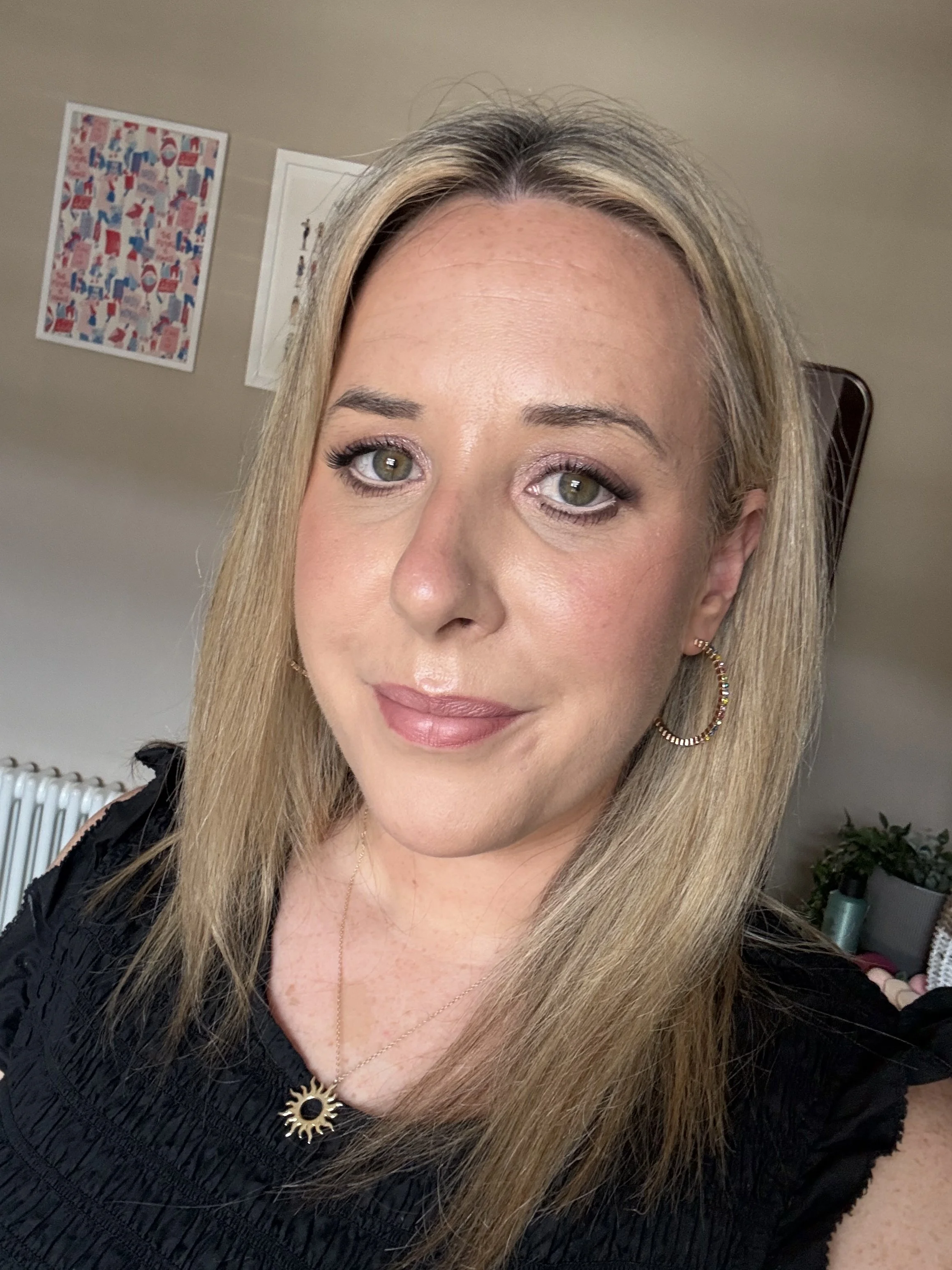 Close-up of a woman with blonde hair, wearing a black top with ruffled details, gold earrings, a gold necklace, and a sun-shaped pendant, smiling slightly with makeup including eye makeup and lipstick, in a room with artwork on the beige wall and a p