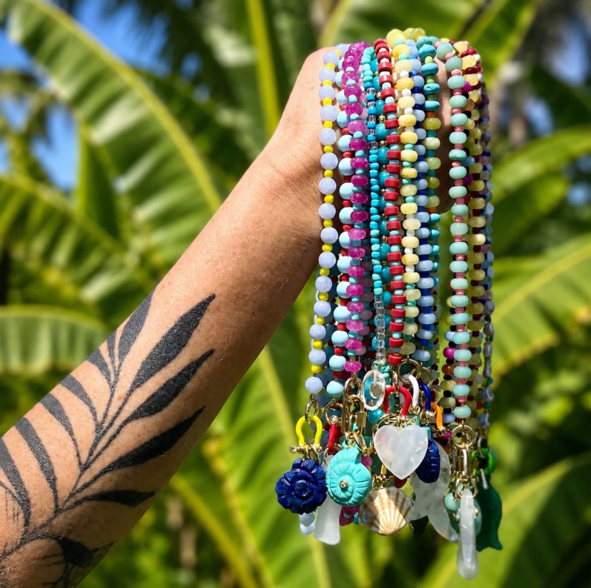Colorful beaded necklaces with charms held up by a tattooed arm against a background of green banana leaves.