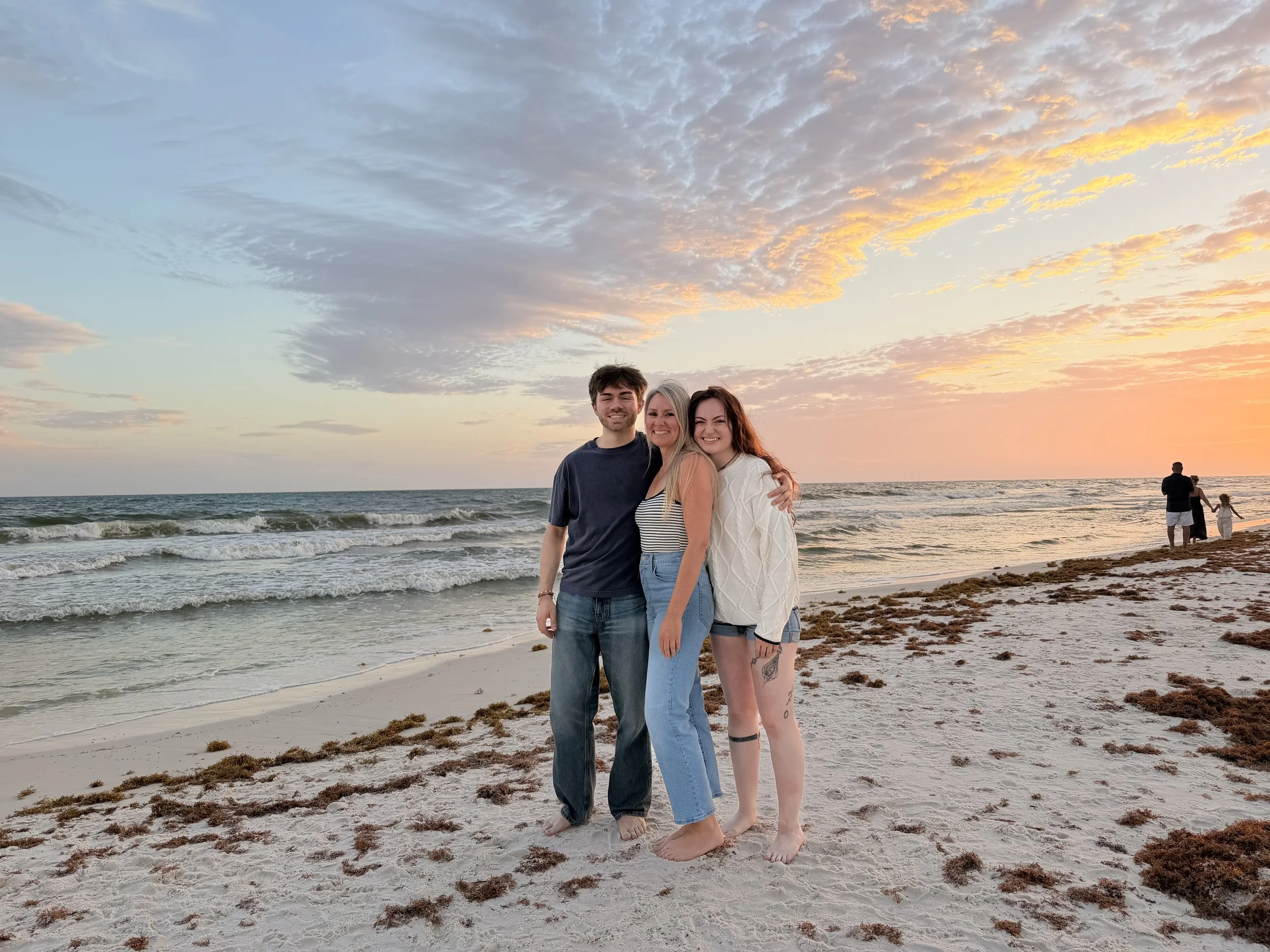 Three friends smiling and hugging on a beach at sunset, with the ocean and sky with clouds in the background.