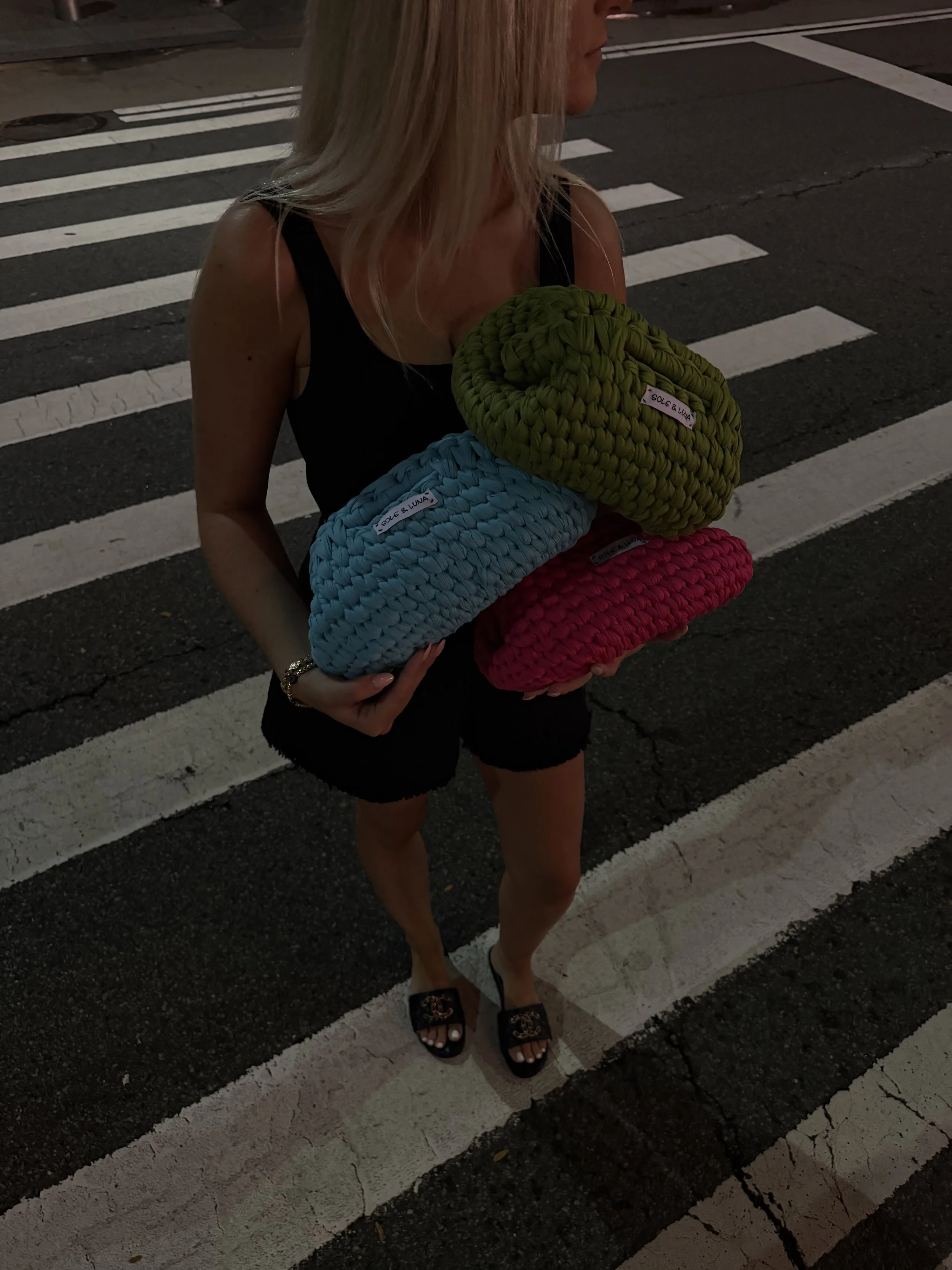 A woman standing on a crosswalk holding three rolled-up blankets or towels in green, blue, and pink colors, with labels that read 'Sole & Luna.' She is wearing a black sleeveless top, black shorts, and black sandals, with a bracelet on her wrist and long blonde hair.