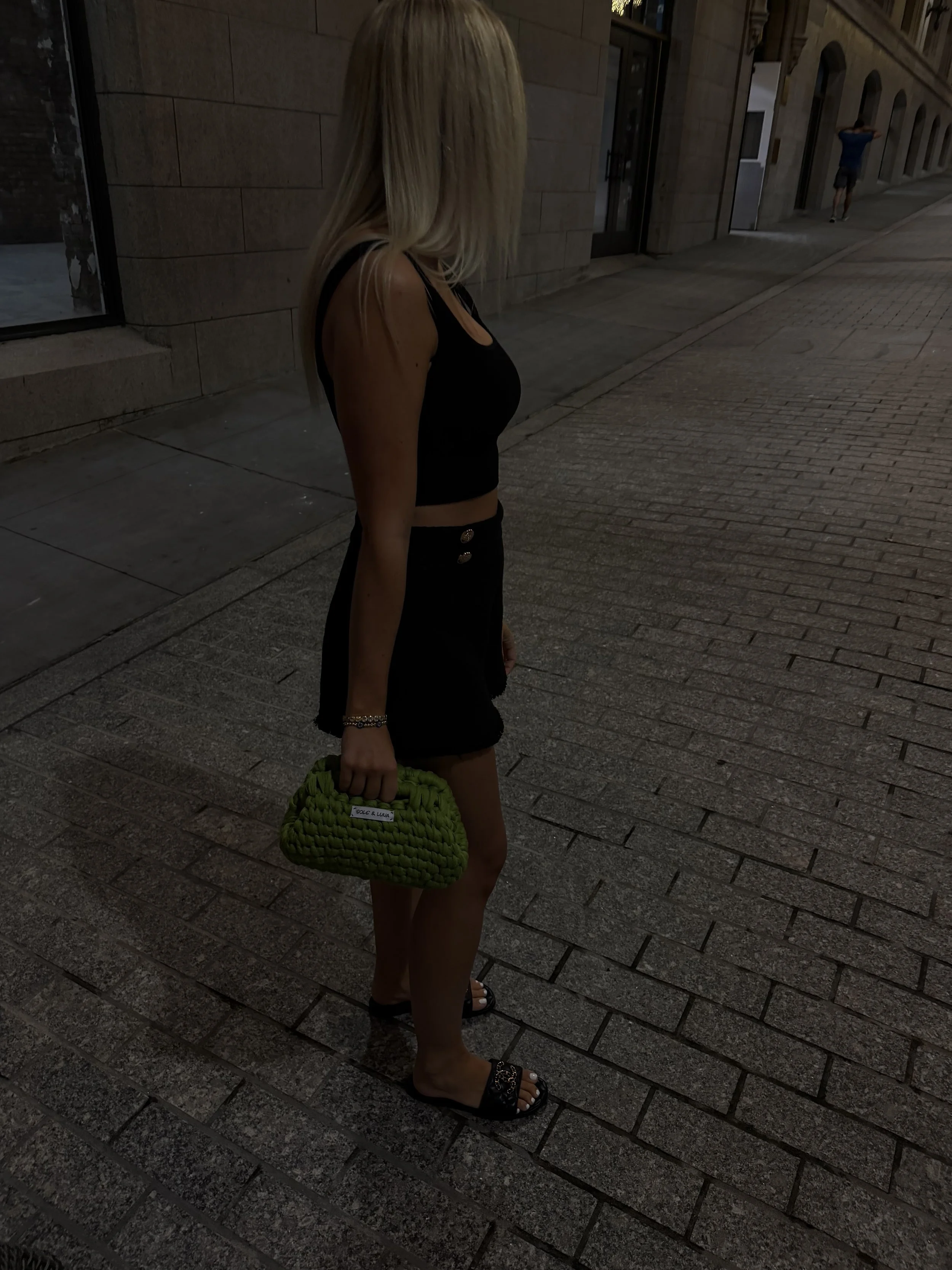 A woman with long blonde hair wearing a black sleeveless crop top, black shorts with gold buttons, and black sandals, holding a green woven handbag, standing on a cobblestone sidewalk at night.