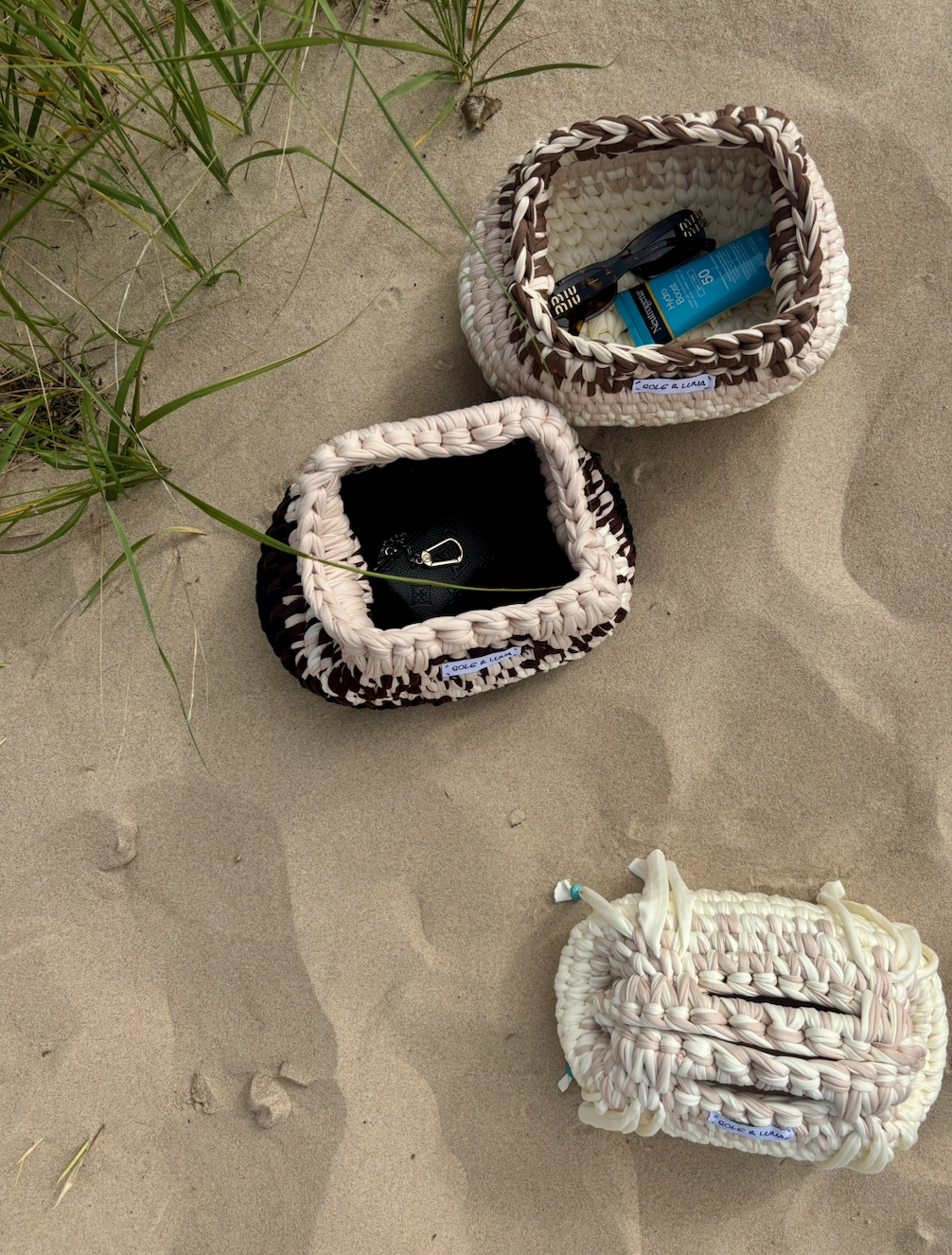 Three woven bags with personal belongings on sandy beach, some with green beach grass nearby.