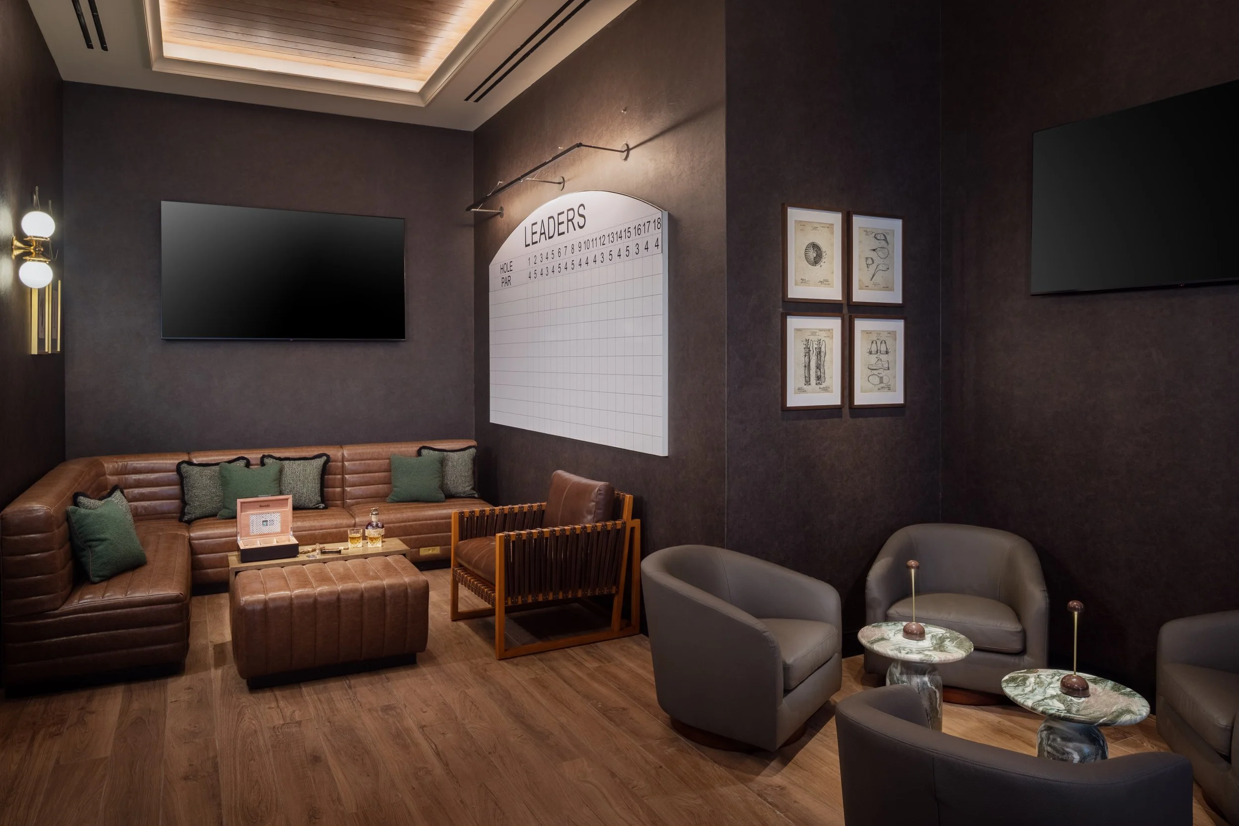 A cozy lounge area with leather couches, green pillows, and small marble-topped tables, featuring a dark wall with framed artwork and a large flat-screen TV.