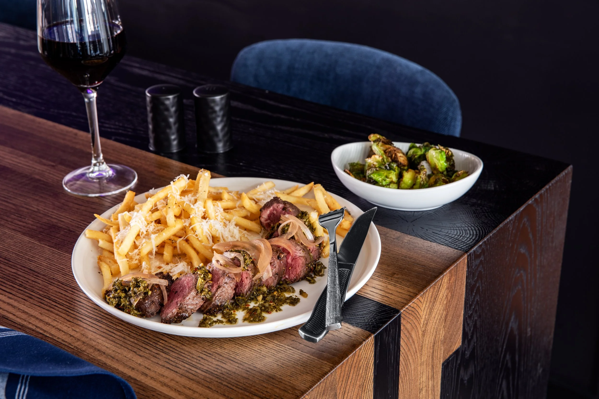 A plate of French fries topped with shredded cheese and cooked steak with herbs, a bowl of cooked Brussels sprouts, and a glass of red wine on a dark wooden table.