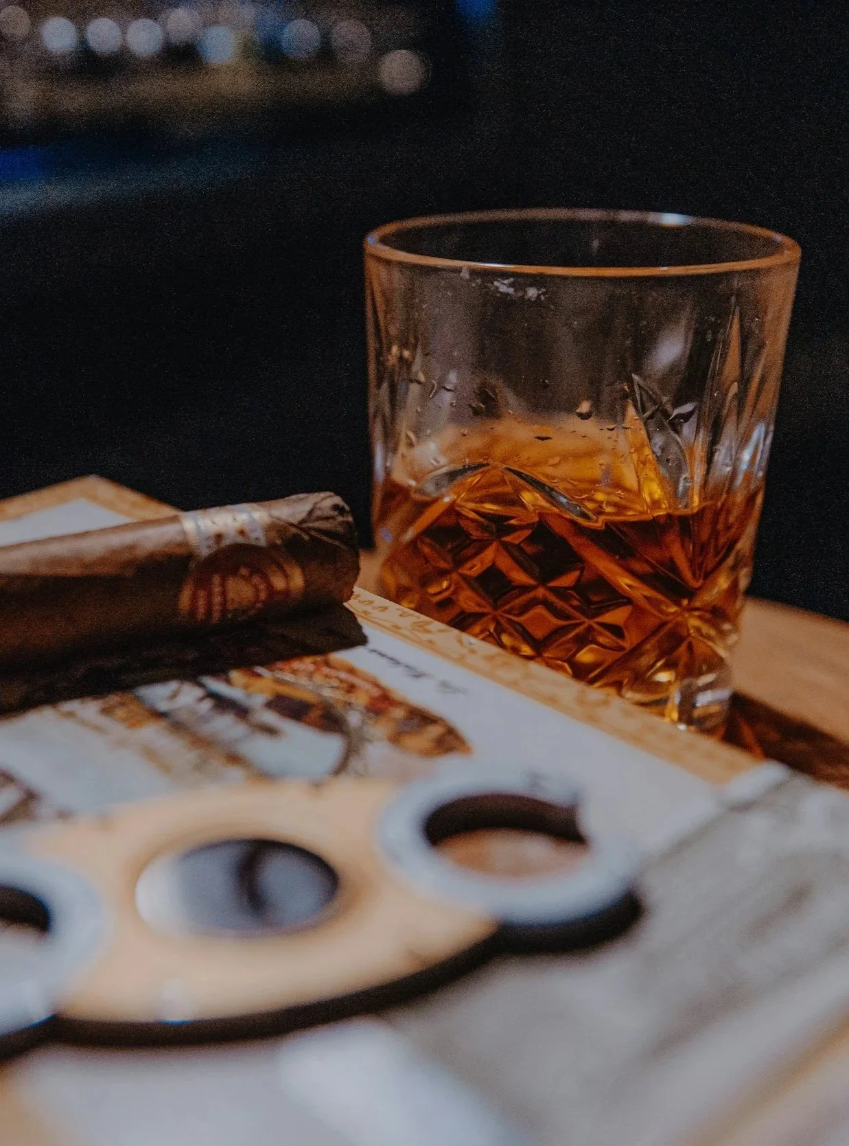 A glass of amber-colored liquor, possibly whiskey or bourbon, is on a table next to a cigar and a playing card with four black circles on it. The background is dark and blurred.