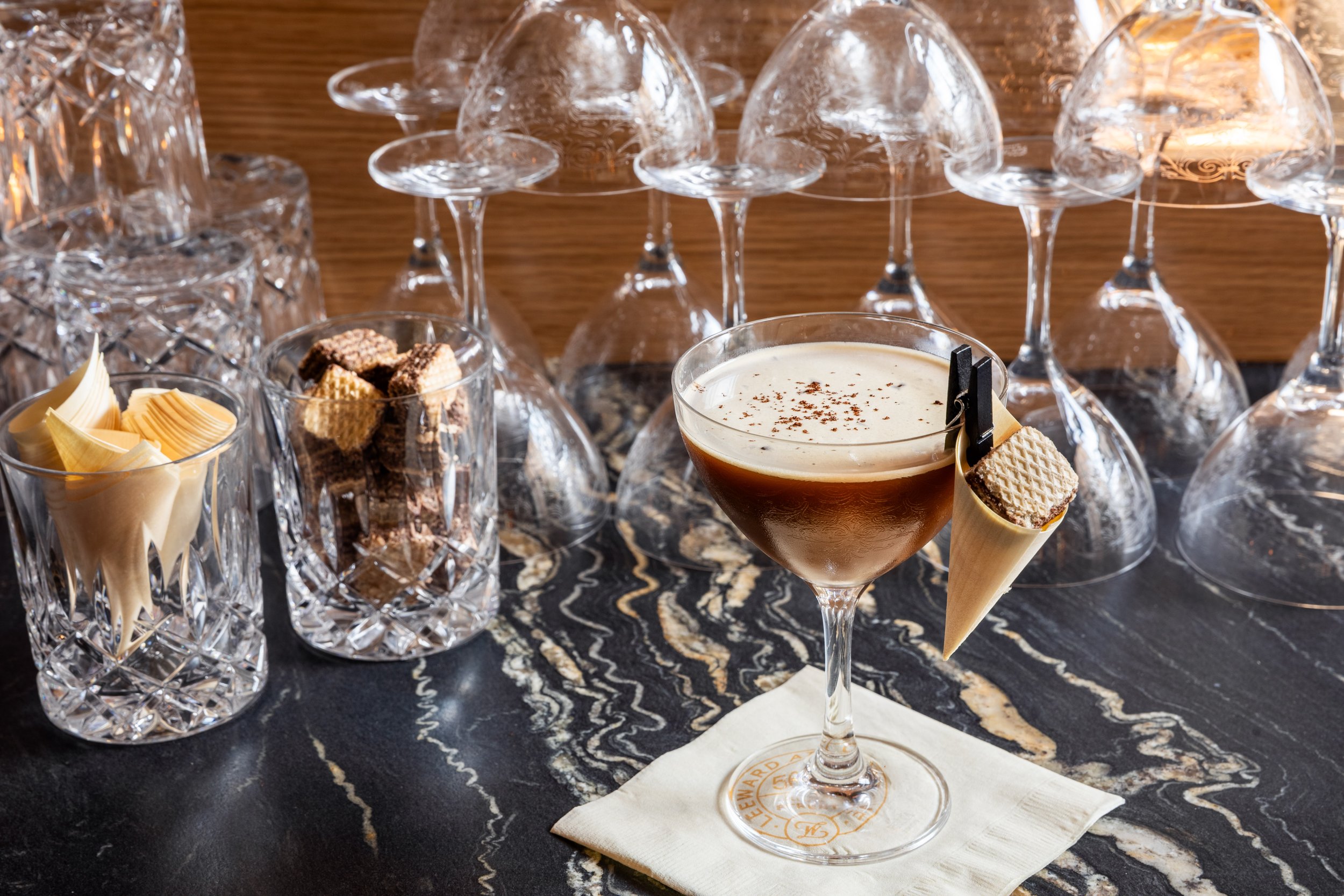 A cocktail in a martini glass with whipped cream and chocolate shavings, surrounded by empty crystal wine glasses and small bowls of snacks on a black marble table.