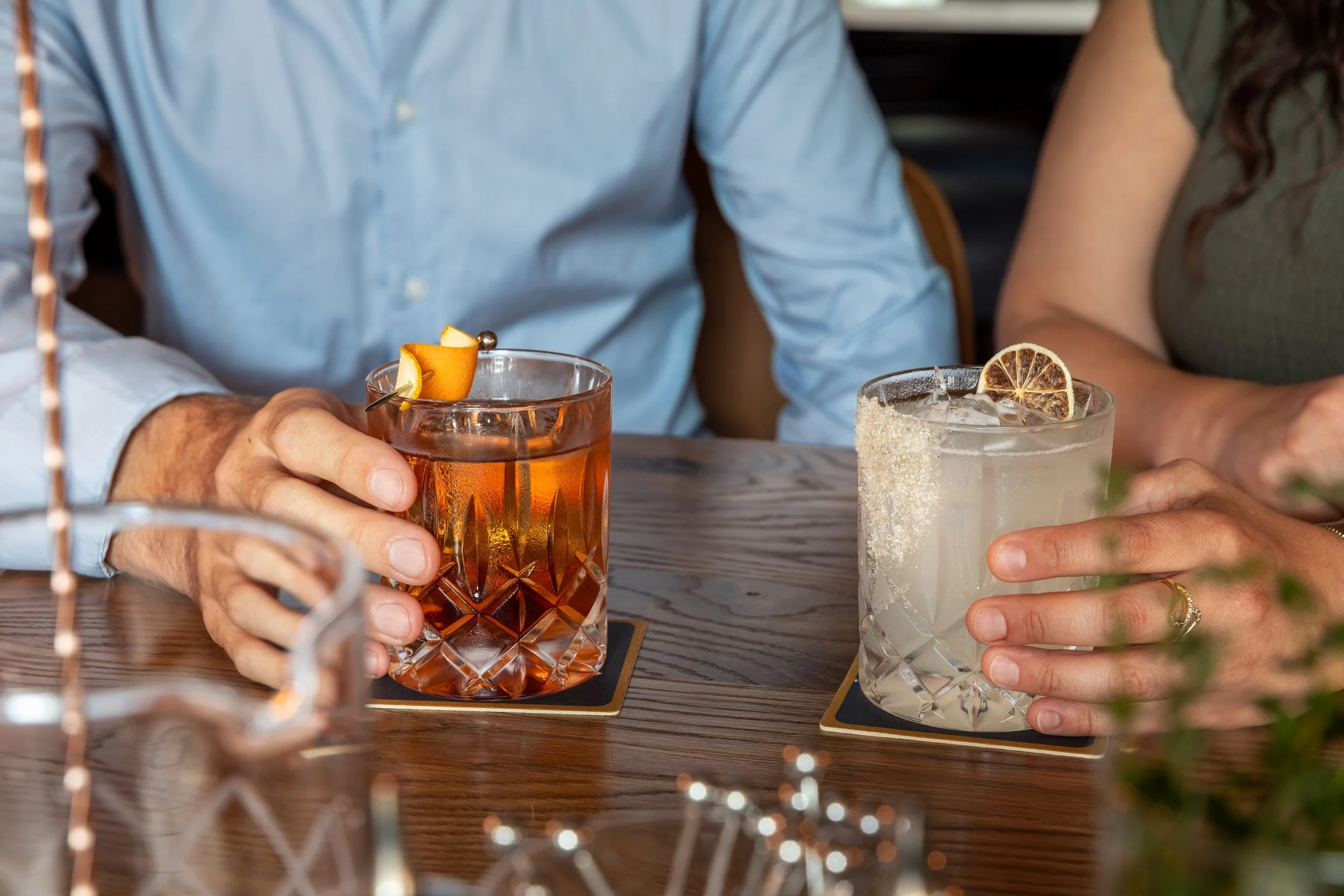 A person's hand holding a glass of iced drink garnished with a lemon slice and orange peel. The person is sitting at a wooden table with other drinks visible, including a cocktail garnished with a lemon slice and sugared rim. The scene appears to be 