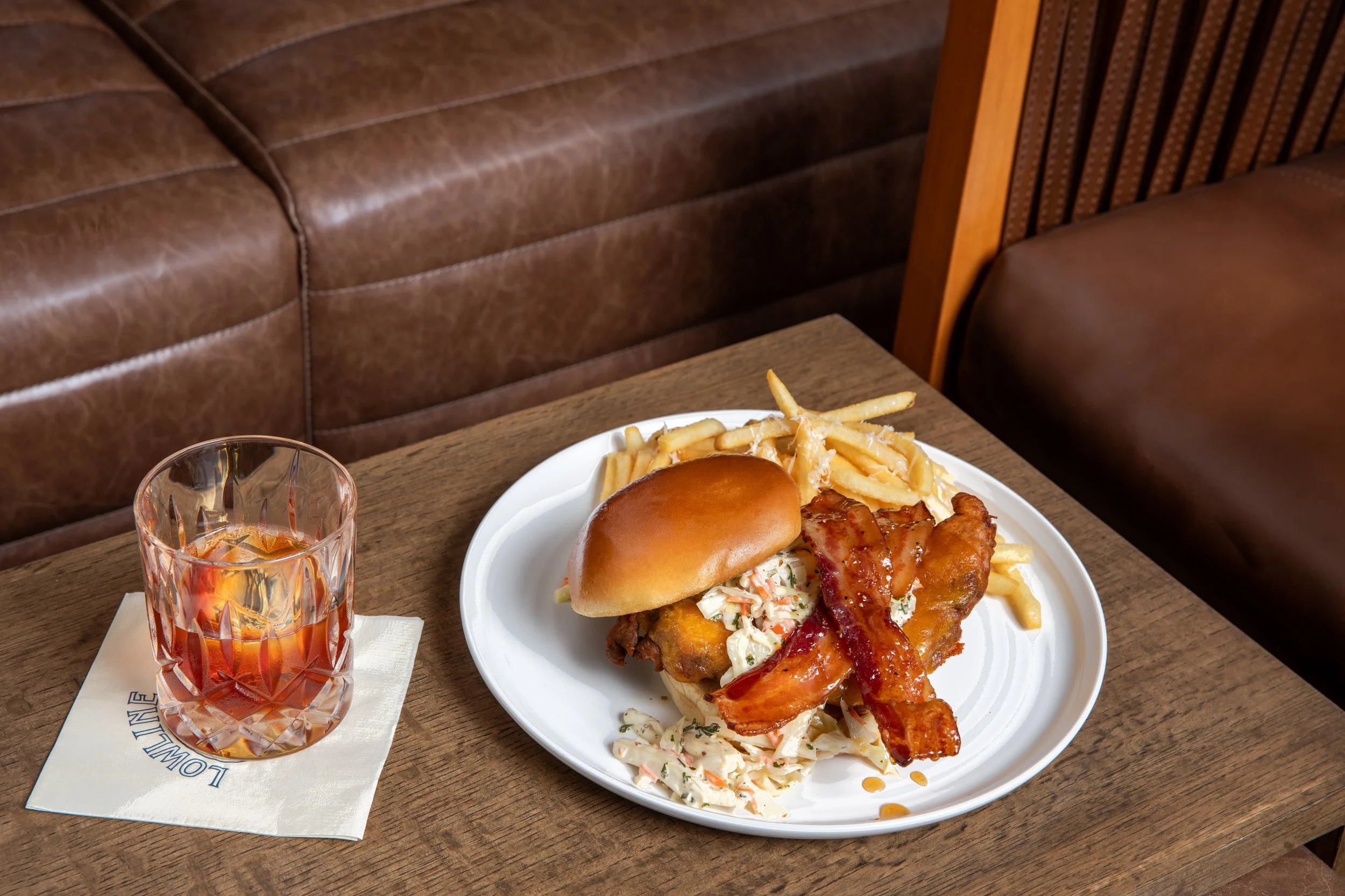 A plate of food with a sandwich, fries, and coleslaw next to a glass of iced tea on a wooden table in a restaurant setting, with brown leather and wooden seating in the background.