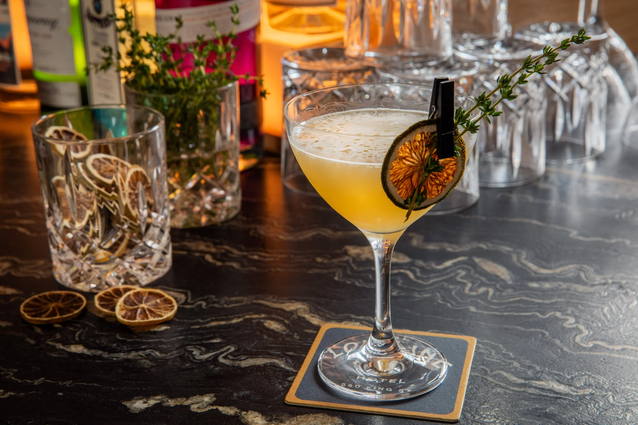 A martini cocktail garnished with a dried lemon slice and a sprig of thyme, placed on a bar counter with dried lemon slices and various glassware and bottles in the background.