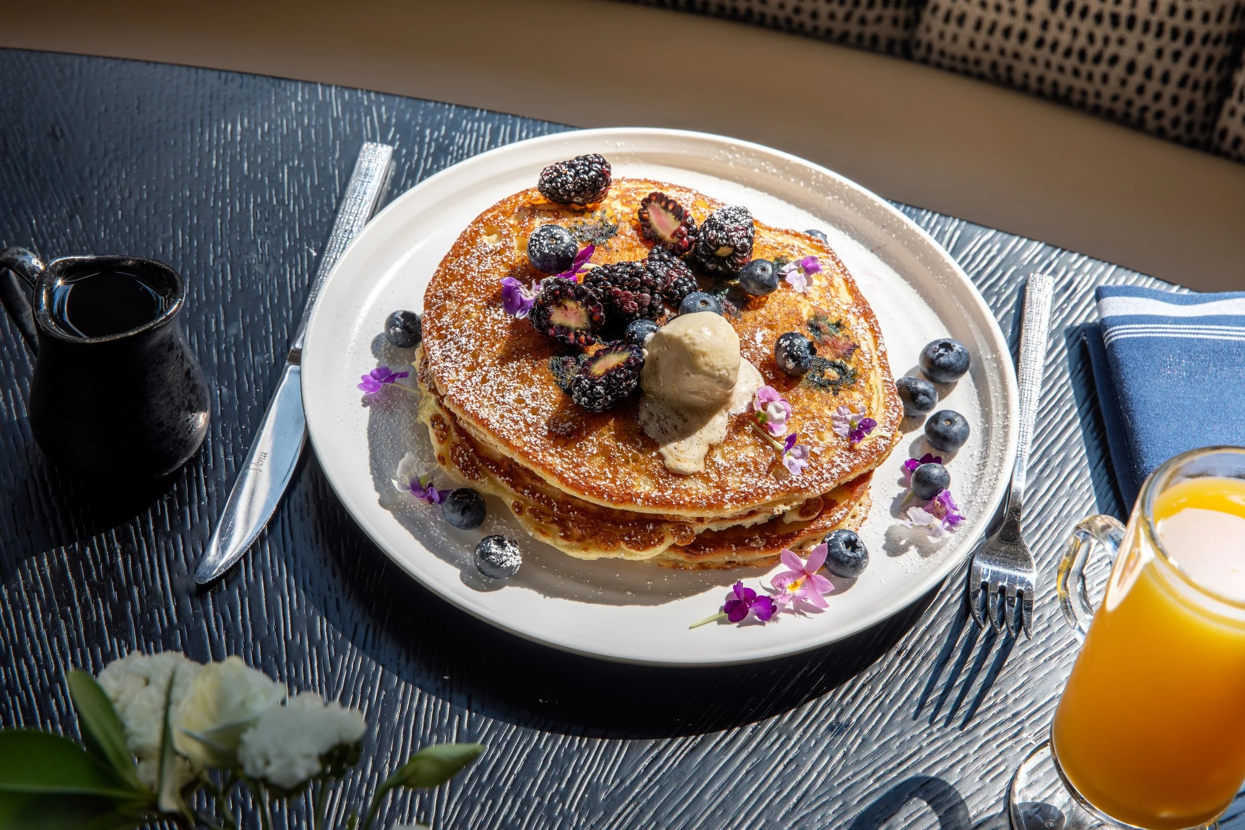 Stack of pancakes topped with blackberries, blueberries, a dollop of butter, and edible flowers on a white plate. There is a small black pitcher of syrup, a blue napkin, and a glass of orange juice on a black textured table.