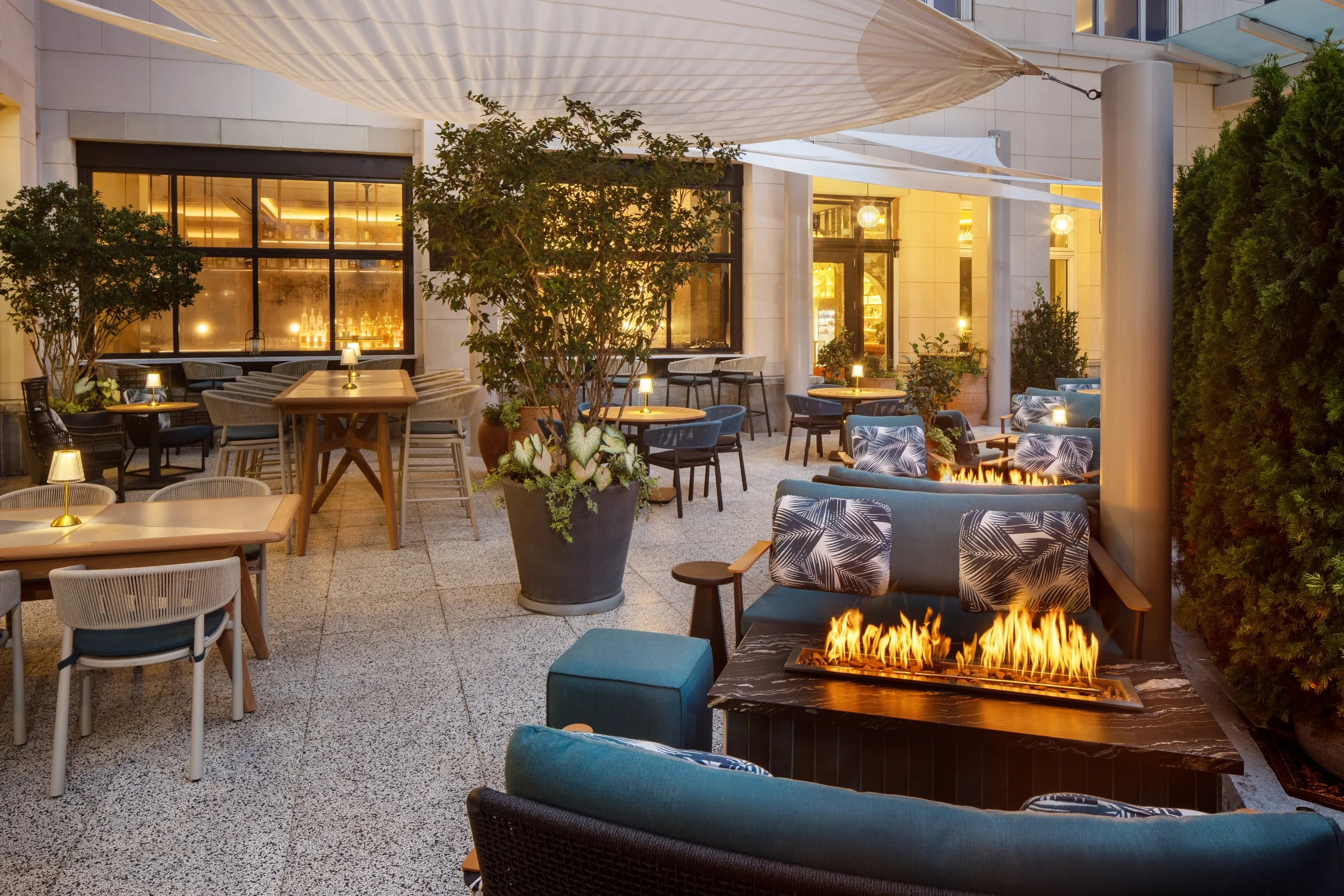 Outdoor patio with seating, tables, and a fire feature, surrounded by plants and buildings, with warm lighting and canopy overhead.