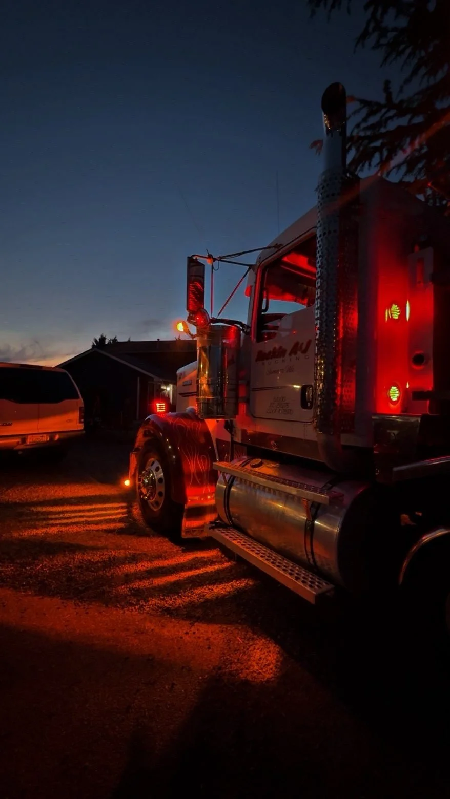 A Kenworth W900 parked outdoors during twilight, with its safety lights illuminated and shadows cast on the ground.