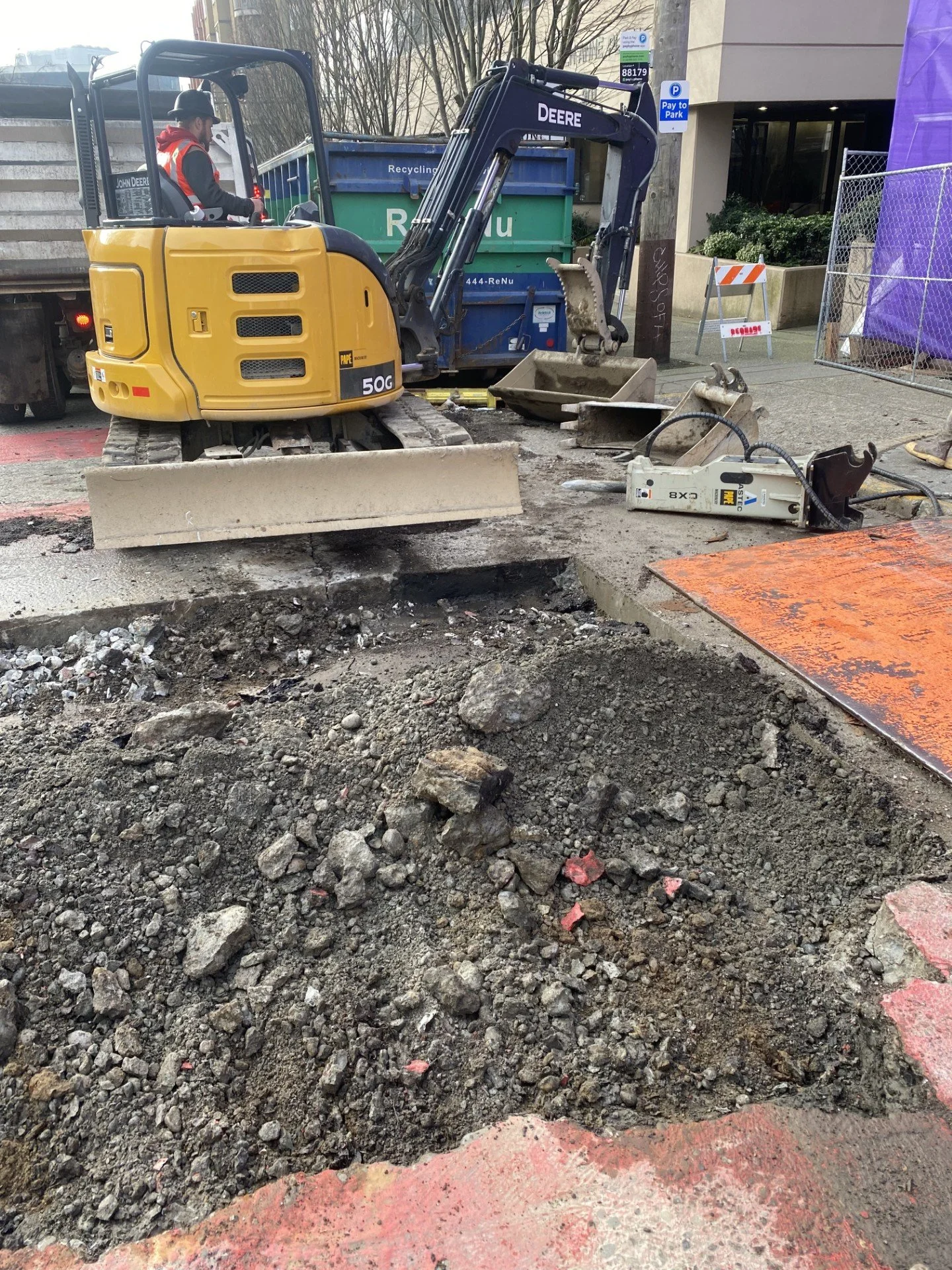 A construction site with a small yellow excavator digging a hole in the street. Construction tools and debris are scattered around, and a worker is in the background operating the equipment. There are construction barriers and fencing, with a green recycling bin nearby.