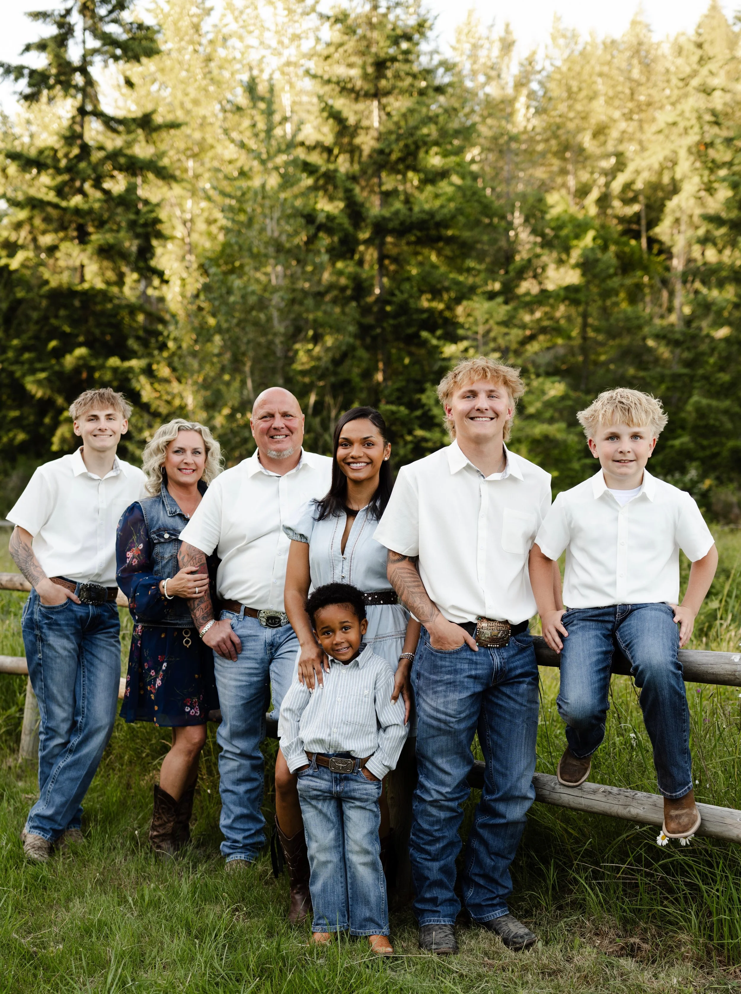 Group of six smiling people standing outdoors on grass, with trees in the background. The group includes adults and children dressed casually.