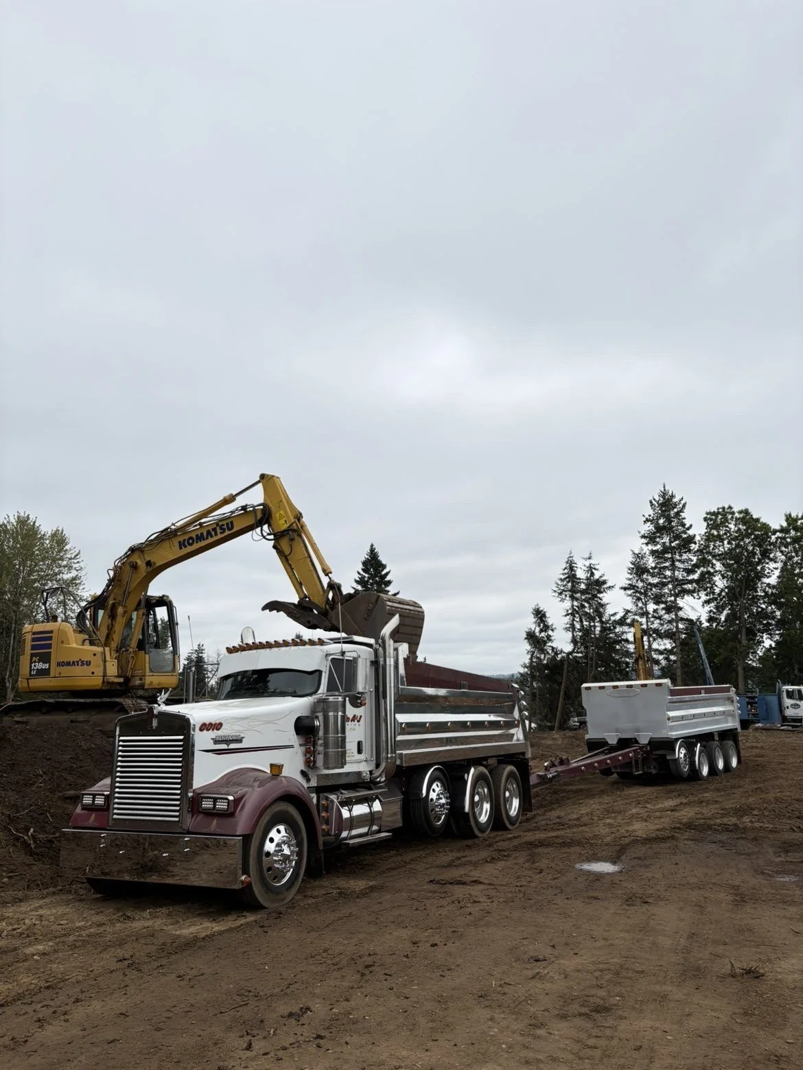 A construction site featuring a yellow Komatsu excavator on an elevated dirt mound and a large white and maroon semi-truck with a trailer parked on dirt ground, with trees and overcast sky in the background.