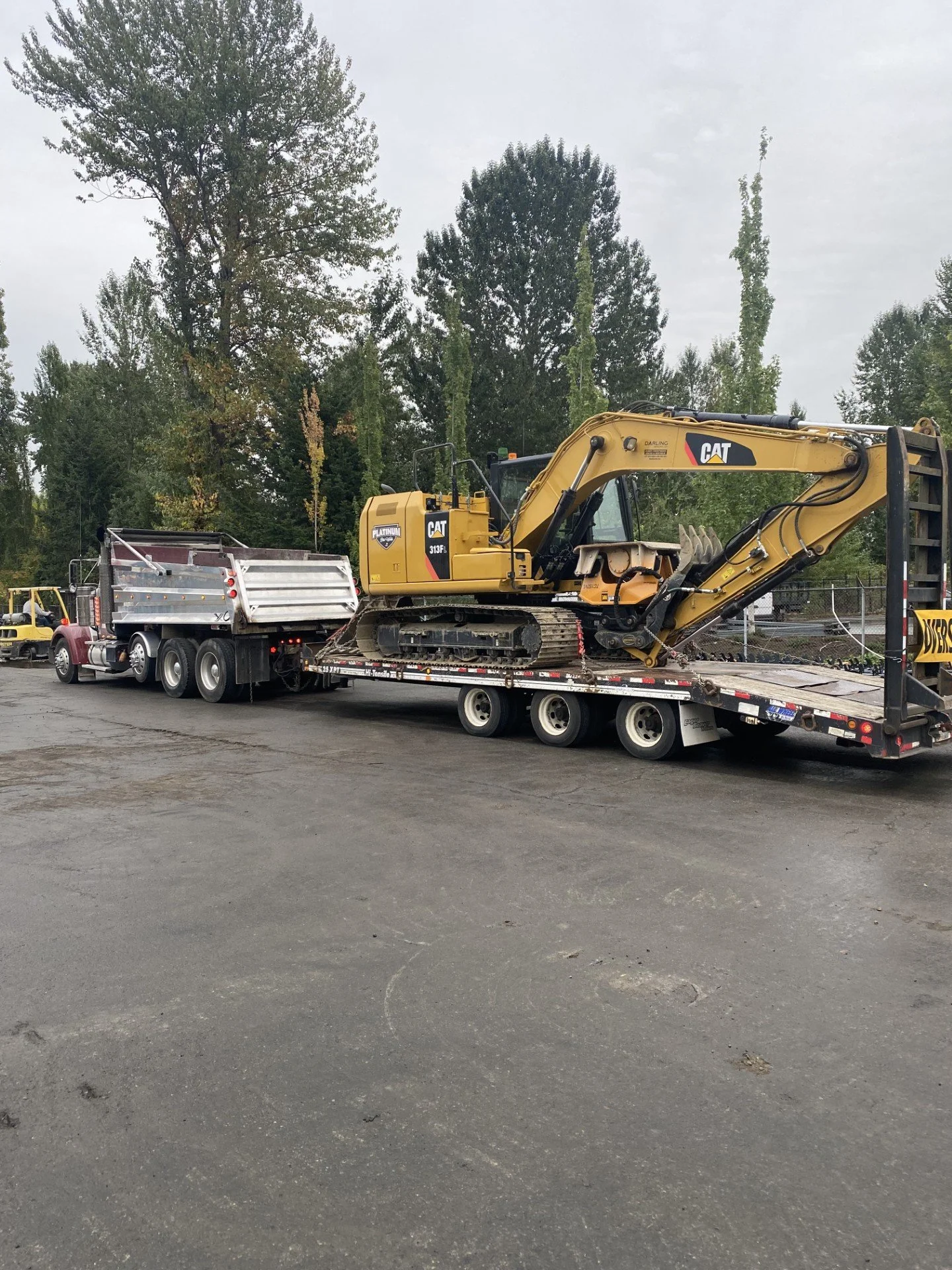 A construction excavator loaded on a flatbed truck in a parking lot with trees in the background.