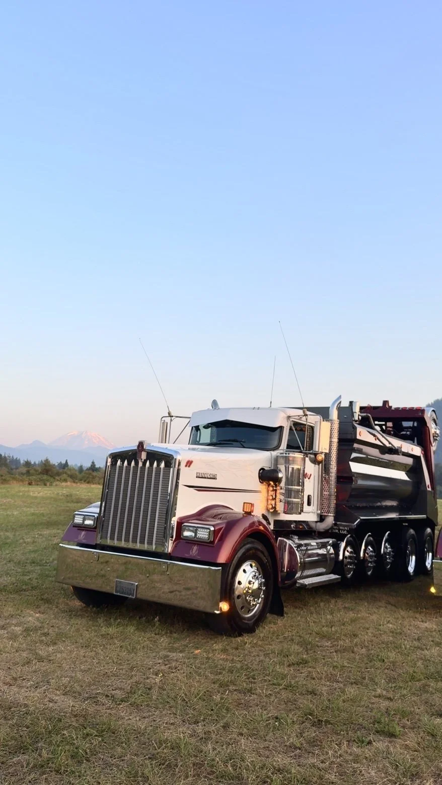 A large white and maroon semi-truck parked on a grassy field with mountains in the background.
