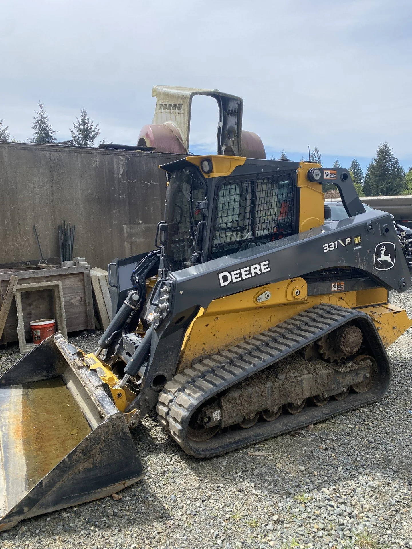 A John Deere mini excavator with black and yellow coloring, tracked wheels, and a front bucket is parked on gravel. It is at a construction site near a concrete wall with trees in the background.