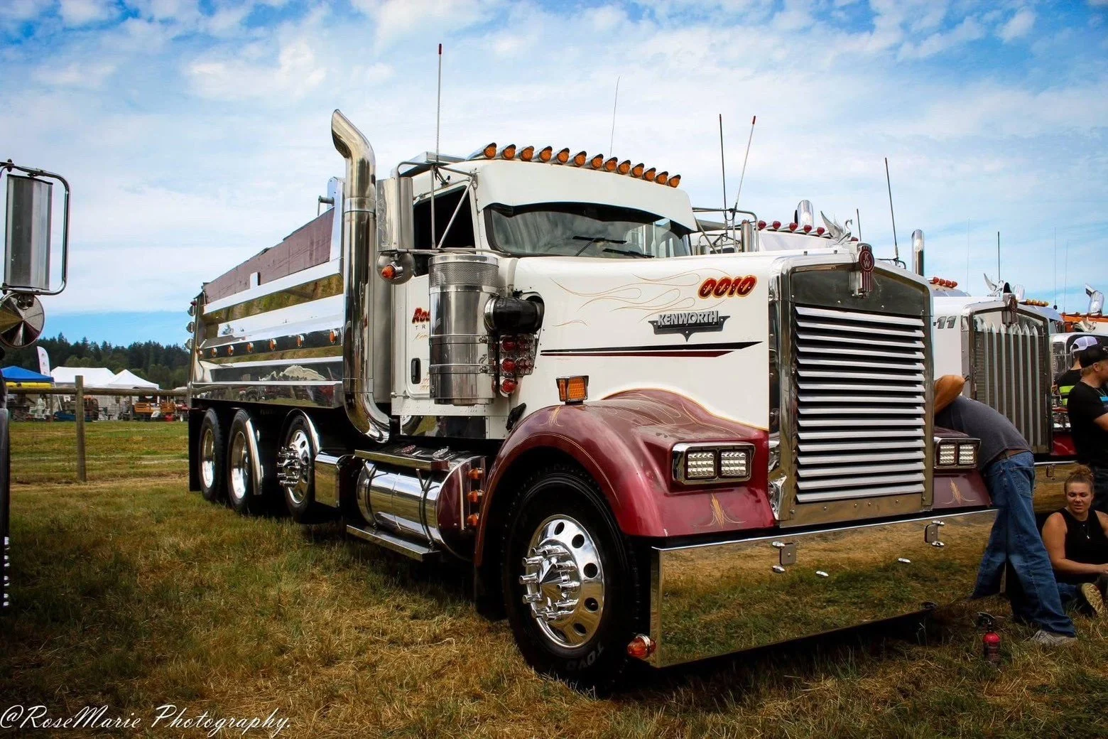 A large vintage Kenworth semi-truck with a white and red color scheme, parked on a grassy field at a truck show. The truck features chrome accents, multiple amber running lights on top, and people gathered around.