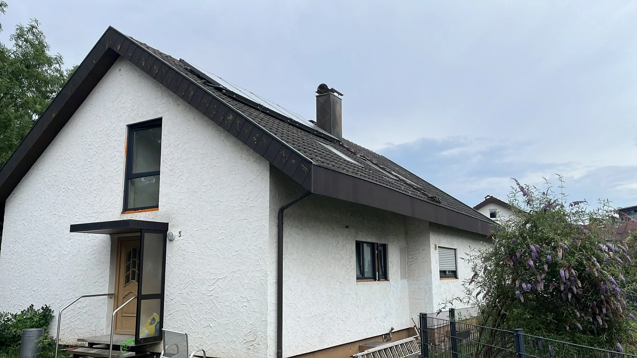 Das Bild zeigt ein weiß gestrichenes Haus mit dunklem Dach. Es hat mehrere Fenster, eines an der oberen Etage und zwei an der unteren. Es gibt einen Hausnummernschild '5' neben der Tür. Im Vordergrund sieht man terrassentürartige Fenster, einen Einga