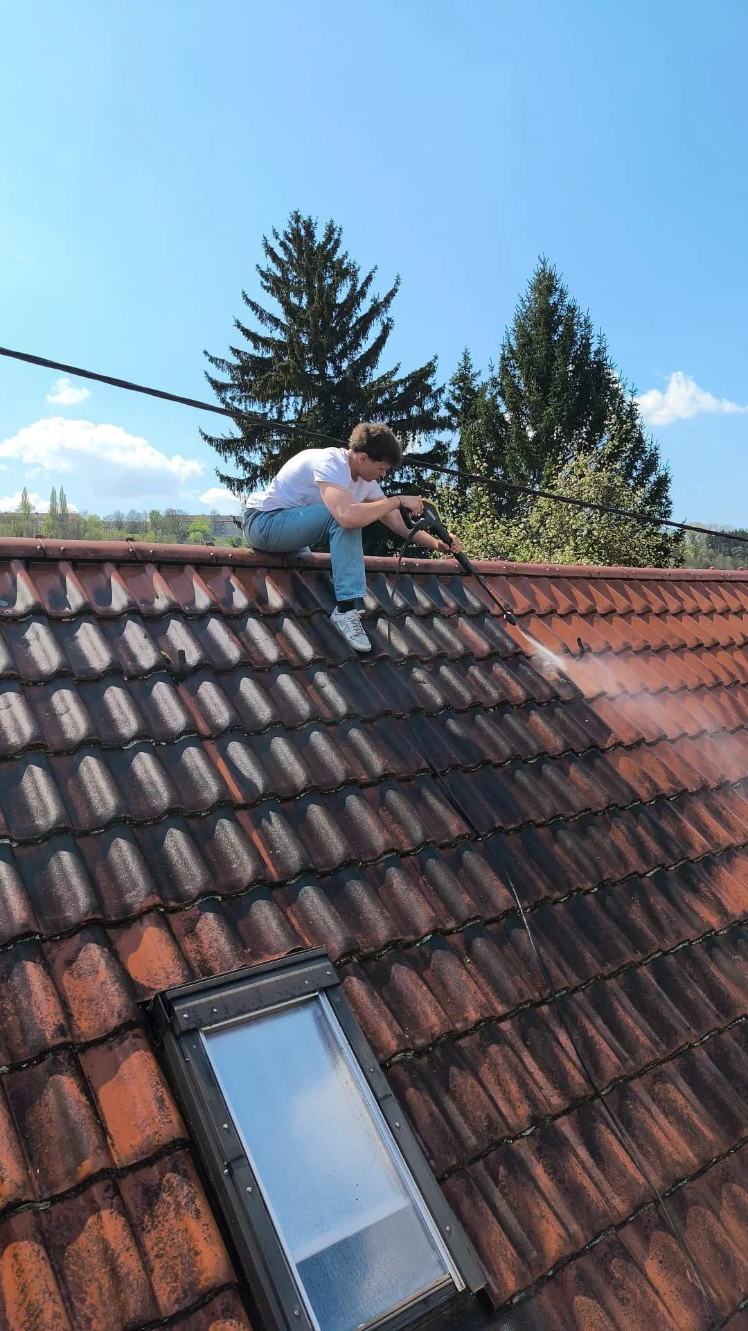 Junge Person reinigt Dachziegel mit Hochdruckreiniger, während die Sonne scheint und die Bäume im Hintergrund zu sehen sind.