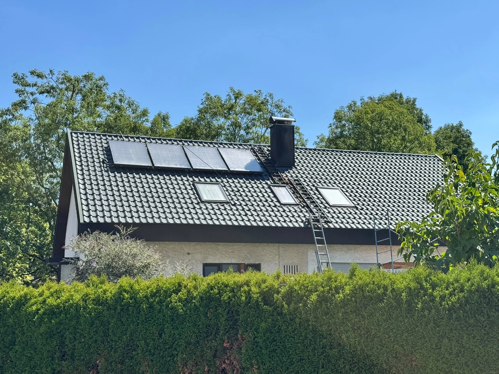 Ein Haus mit Solarpanelen auf dem Dach, umgeben von Bäumen und Sträuchern unter klarem blauen Himmel.