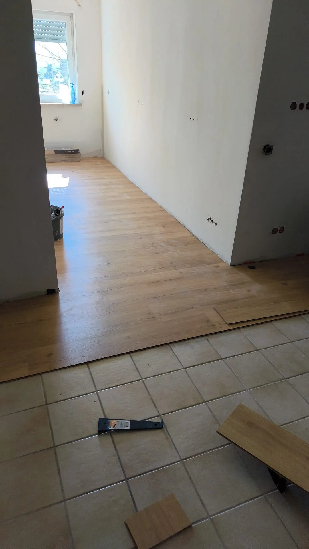 Raum im Umbau mit halb verlegtem Holzboden, noch unvollendeter Wand und einem Fenster mit Blick nach draußen, im Vordergrund Fliese, Werkzeug und Holzplatten.