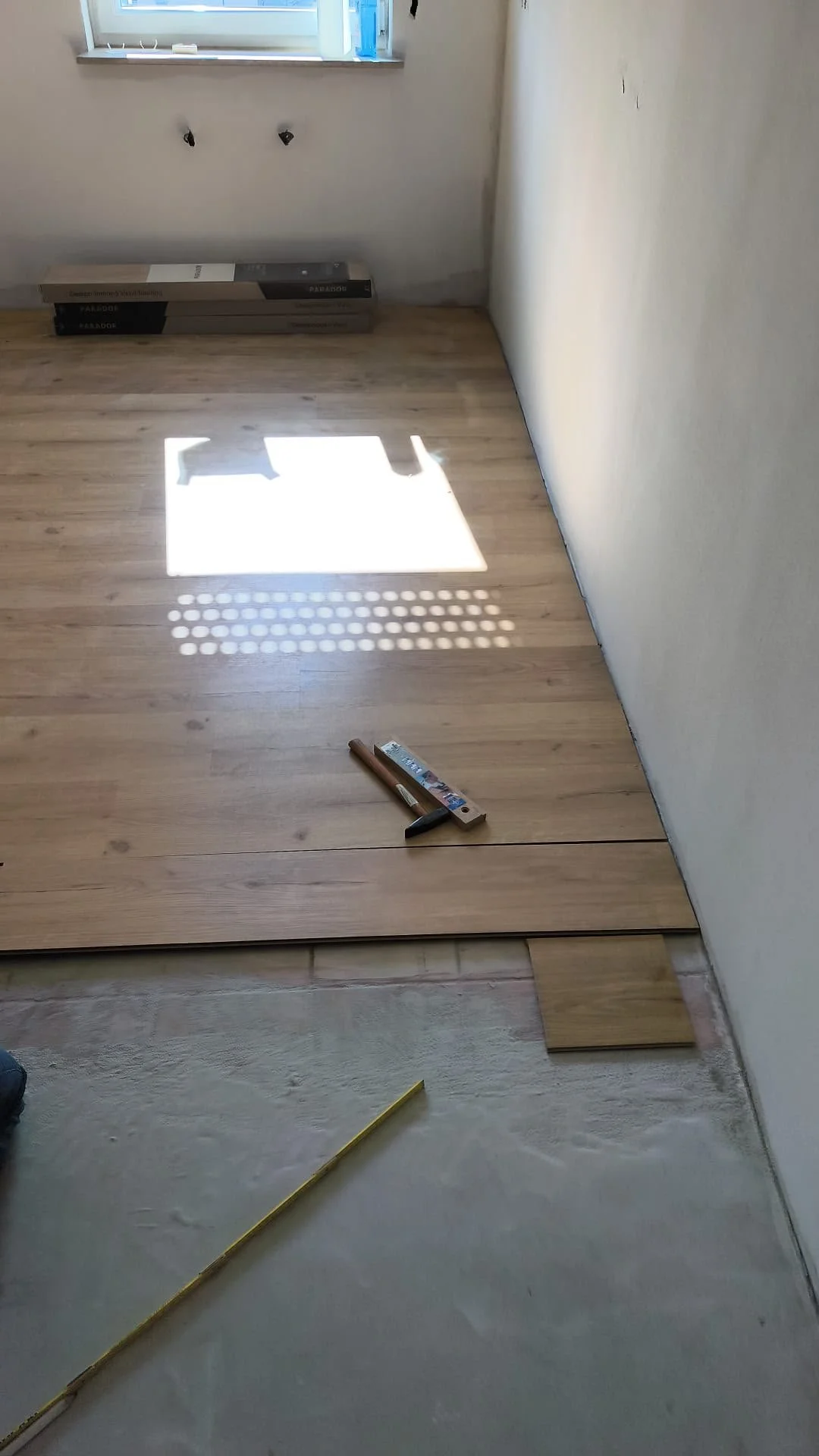 Ein Raum, in dem Parkettbretter verlegt werden, mit Werkzeugen und Kisten im Hintergrund, Sonnenlicht durch das Fenster fällt auf den Boden.