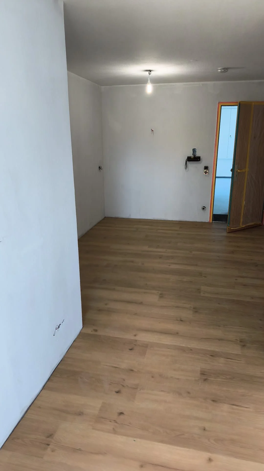 Ein Raum in der Renovierung mit weißen Wänden, Holzfußboden und einer offenen Tür, die zu einem anderen Raum führt.