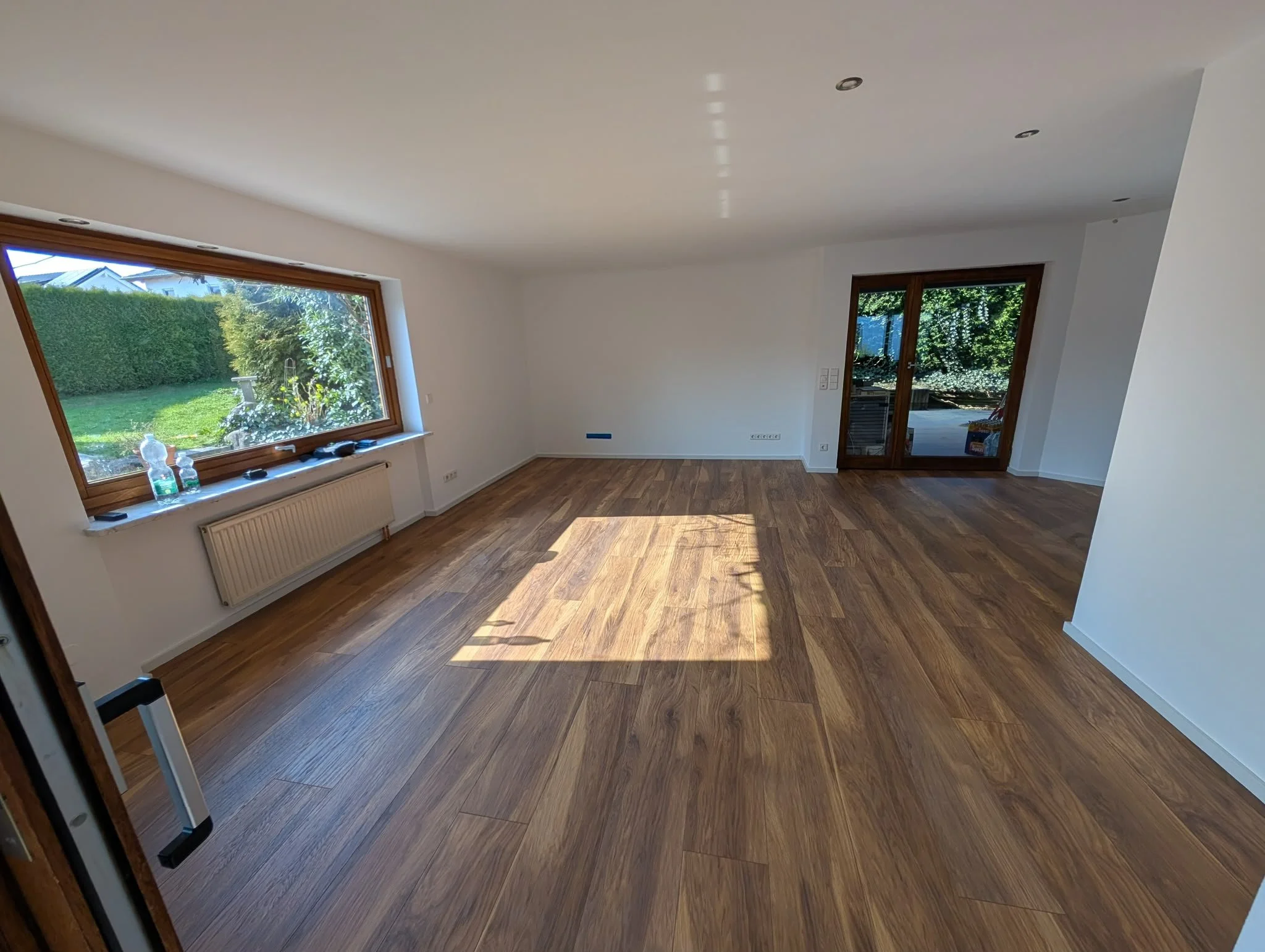 Leeres Wohnzimmer mit Holzfußboden, großen Fenstern und einer Balkontür, Sonne scheint durch die Fenster.
