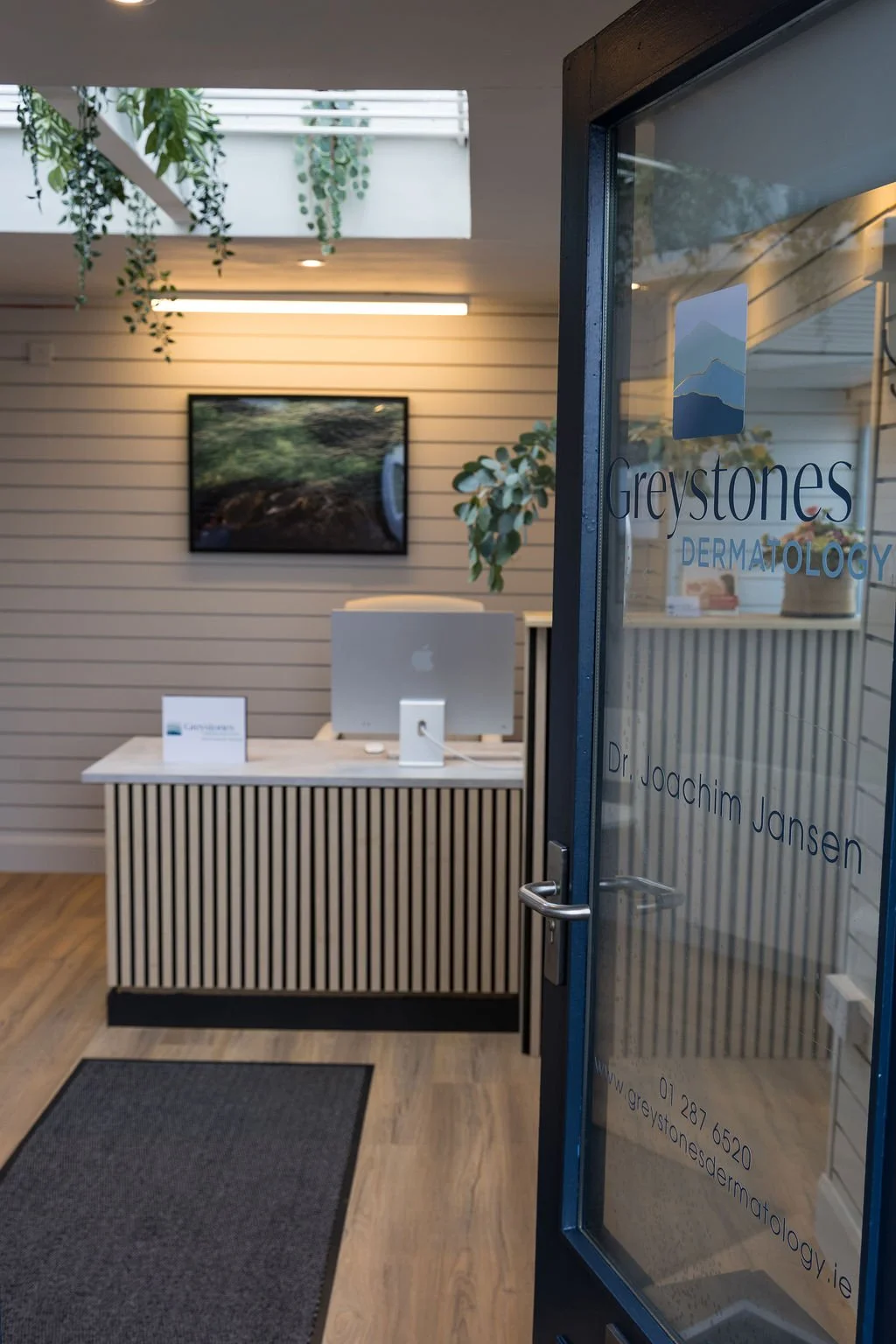 Reception area of Greystones Dermatology with a reception desk, computer, wall-mounted TV, and plants in a well-lit space.