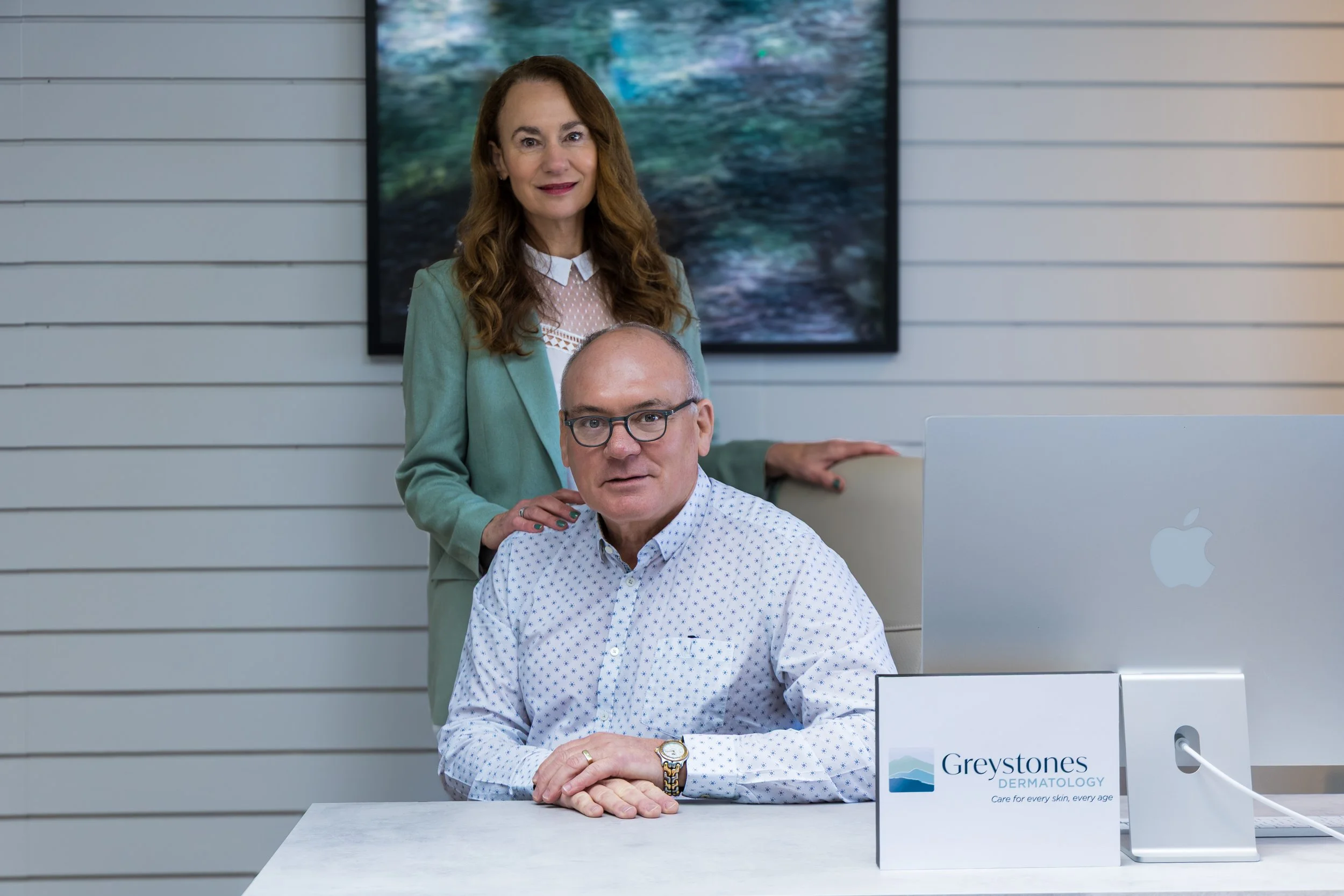 A woman with long curly brown hair, wearing a light green blazer and a lace top, standing behind a seated man with glasses and a patterned white shirt, in an office setting with a logo for Greystones Dermatology on the desk.